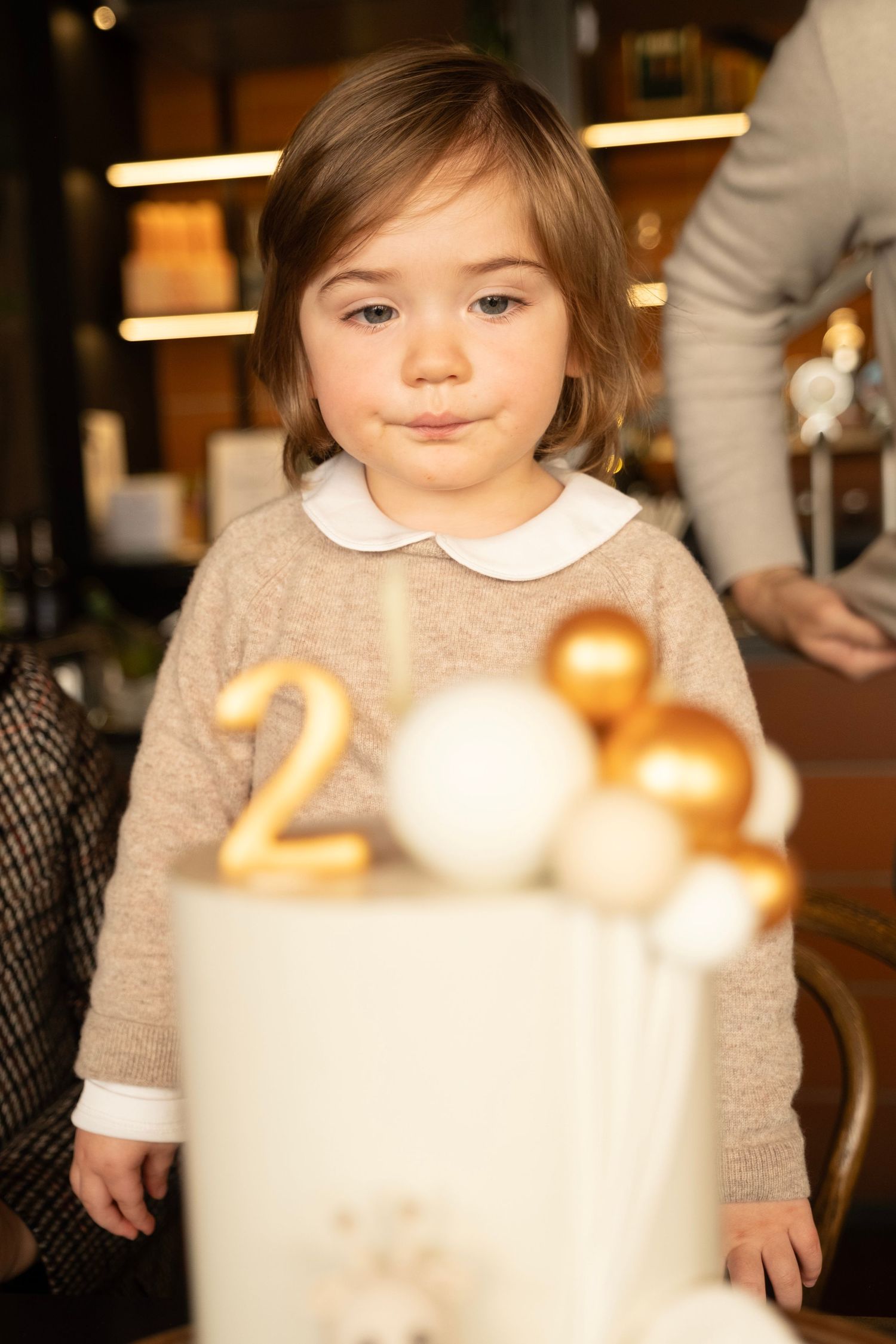 A toddler in a beige sweater with white collar stands near a decorated birthday cake with gold number candles. auckland