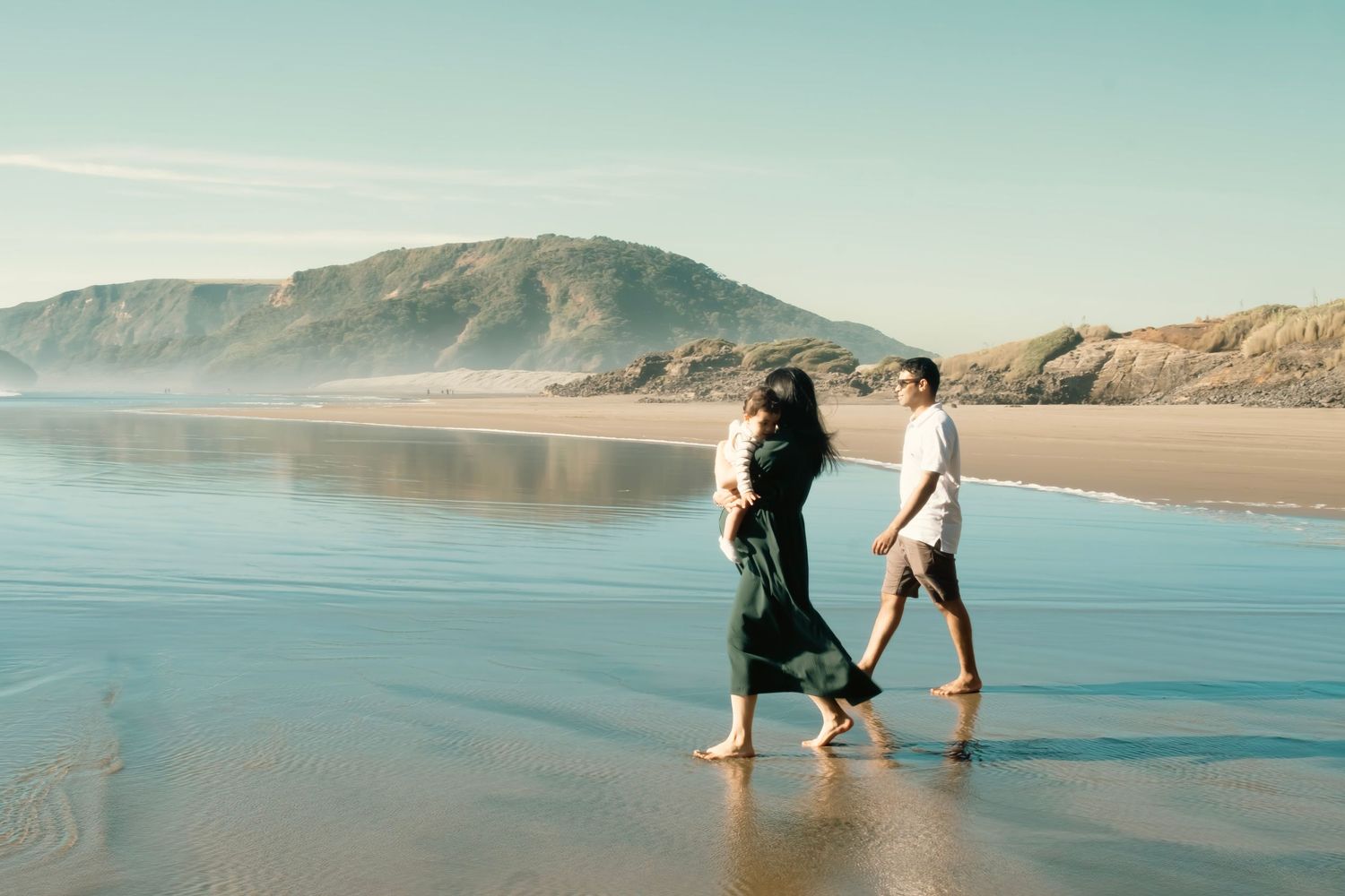 People and dog walking along a serene ocean shoreline with mountains visible in distance during sunset. maternity auckland bethells beach