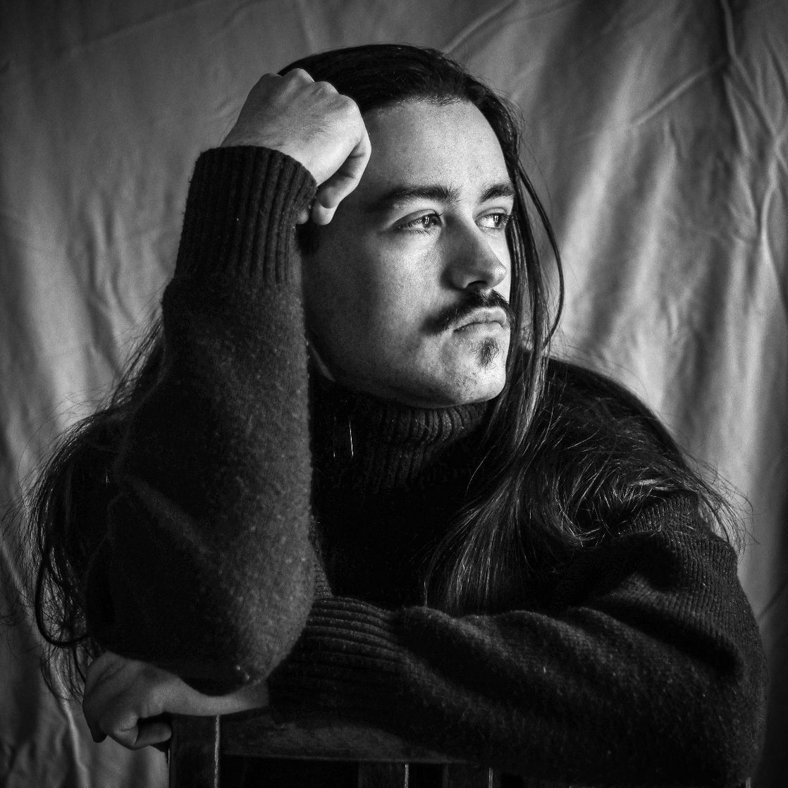 Dramatic black and white portrait of person with long hair and mustache wearing dark sweater in moody lighting.