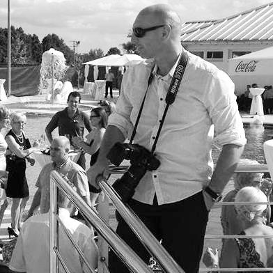 A photographer in a white shirt with sunglasses and camera strap looks out over an outdoor event space in black and white.