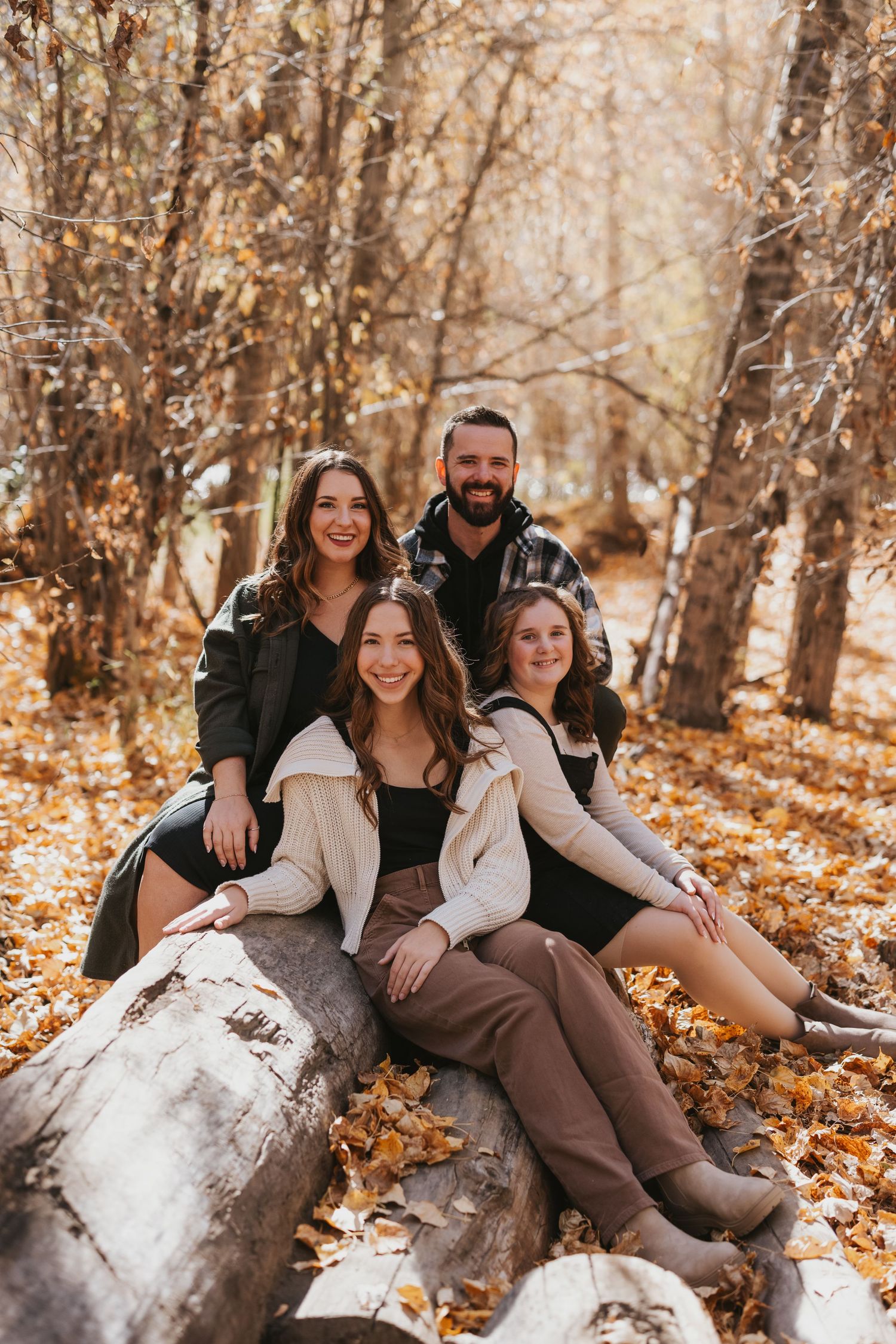 Four people sit together on fallen logs in an autumn forest surrounded by golden leaves and trees.
