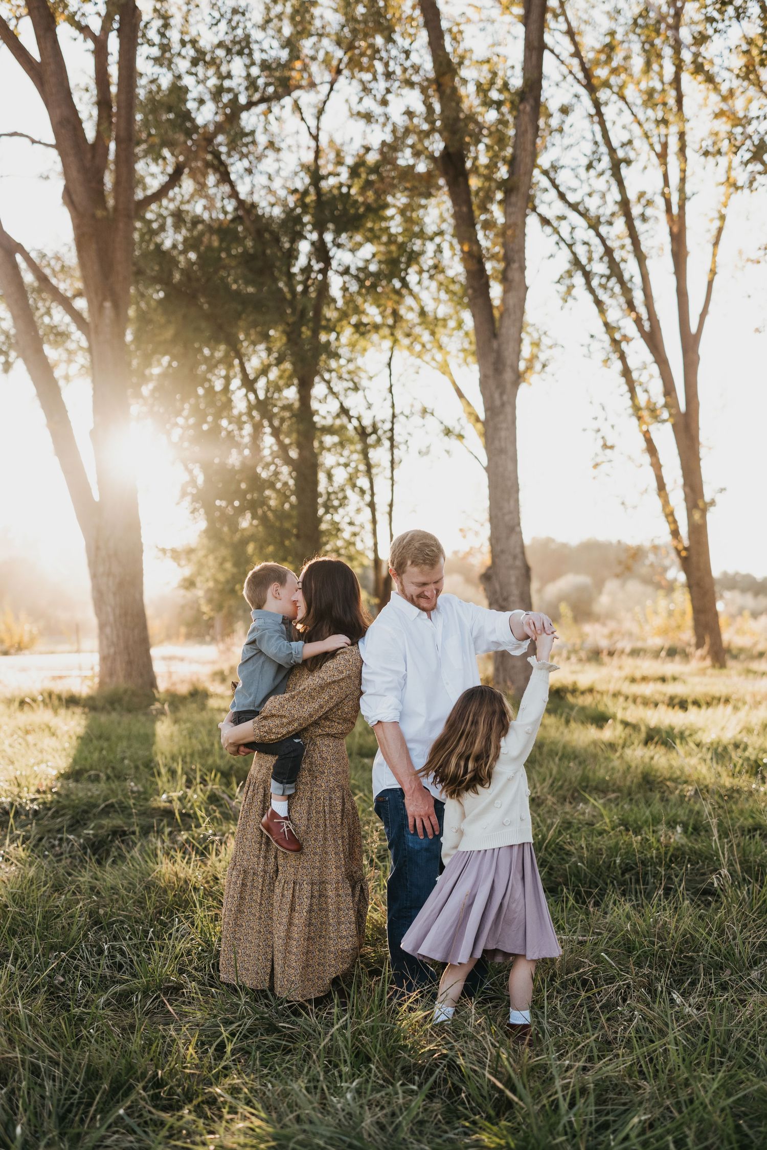 A family stands together in a grassy field during golden hour sunlight with tall trees in the background creating a warm glow.