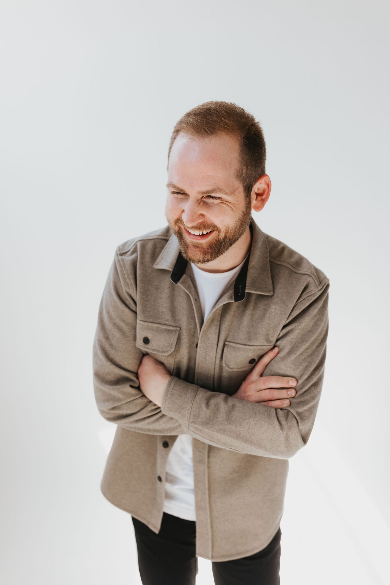 Candid portrait of someone wearing a beige button-up jacket over a white t-shirt, laughing with arms crossed.