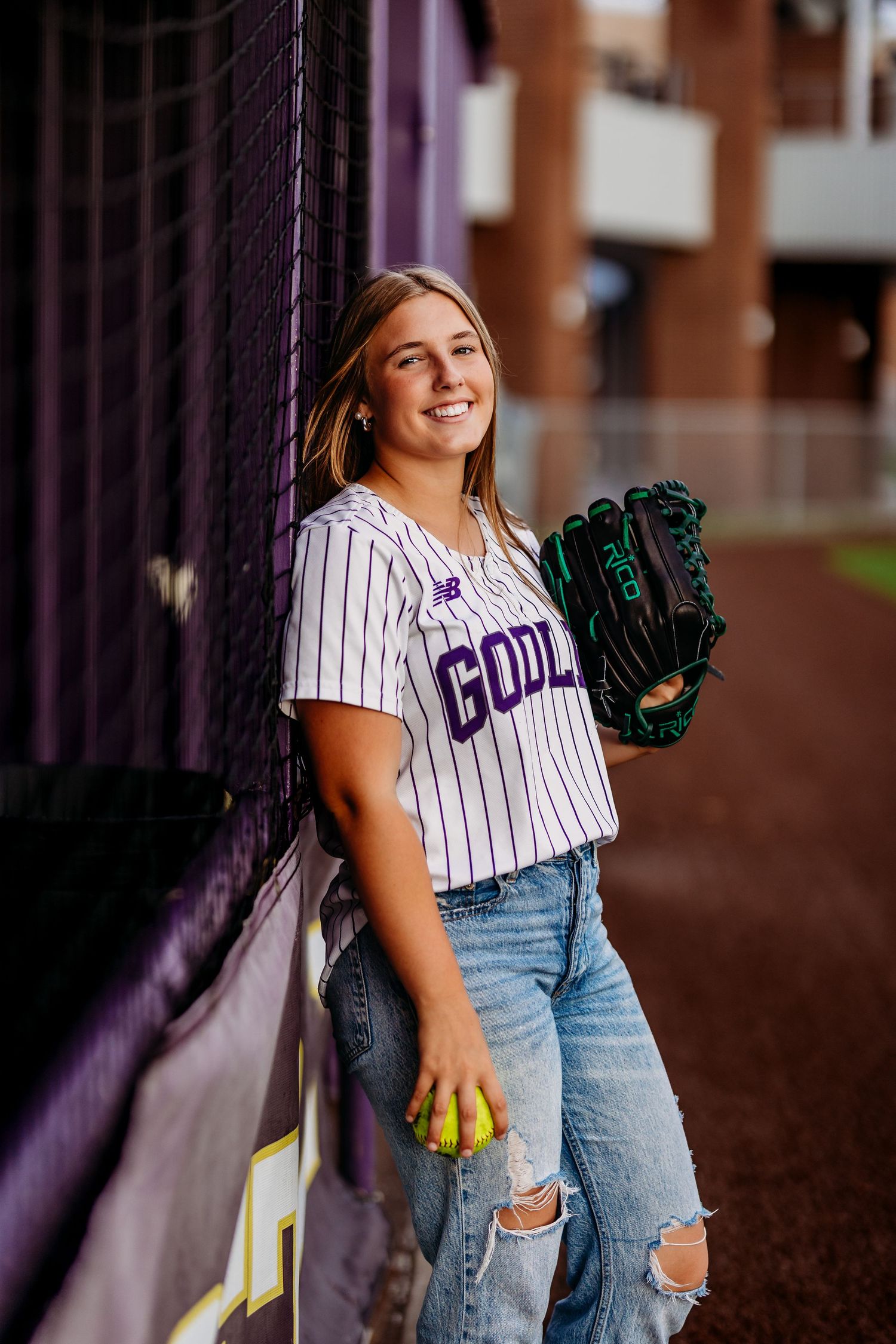 Smiling softball player in Godley pinstripe jersey holding glove and ball, leaning against fence outdoors.