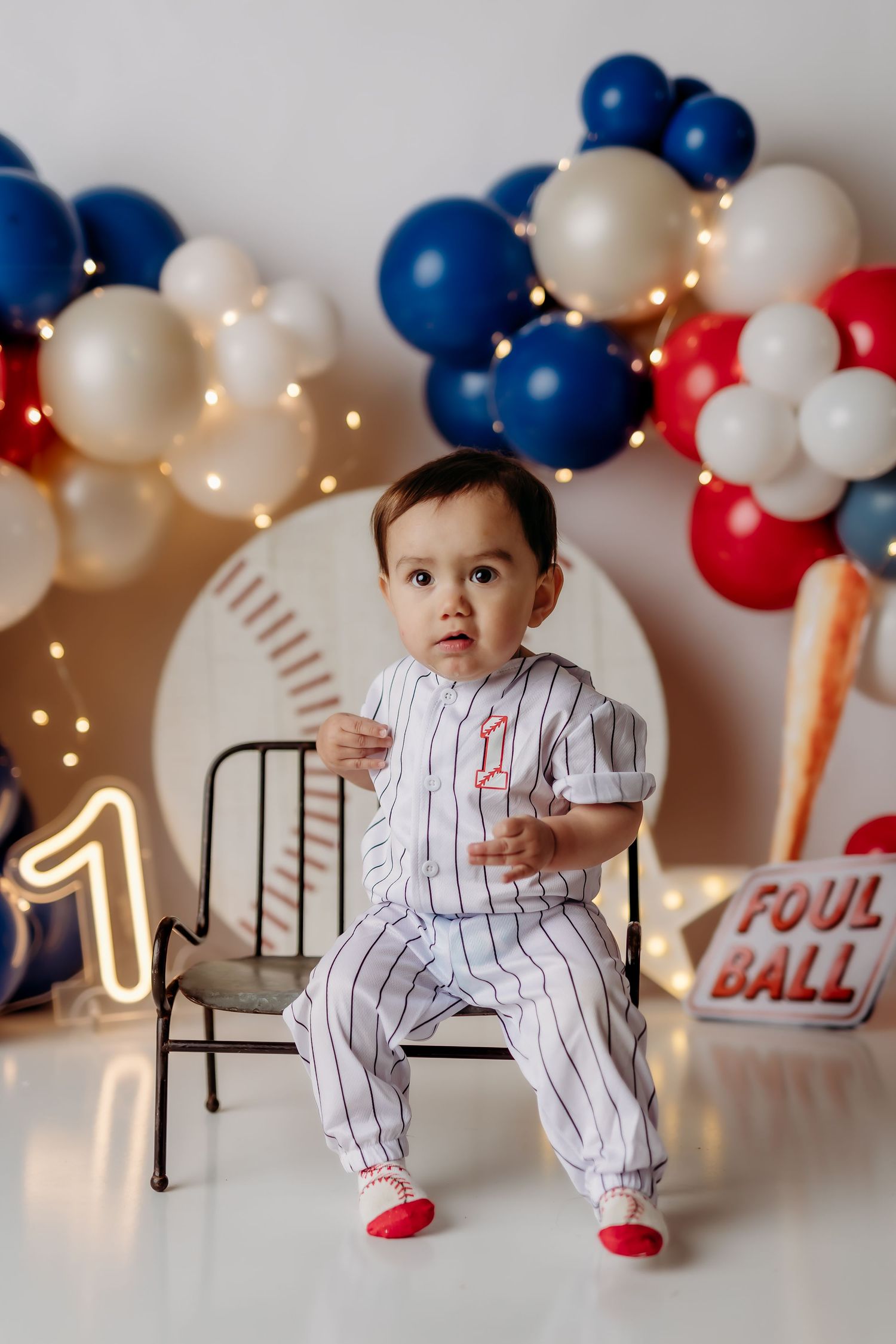 Baby in baseball uniform sits on chair with red, white, and blue balloons for a first birthday celebration.