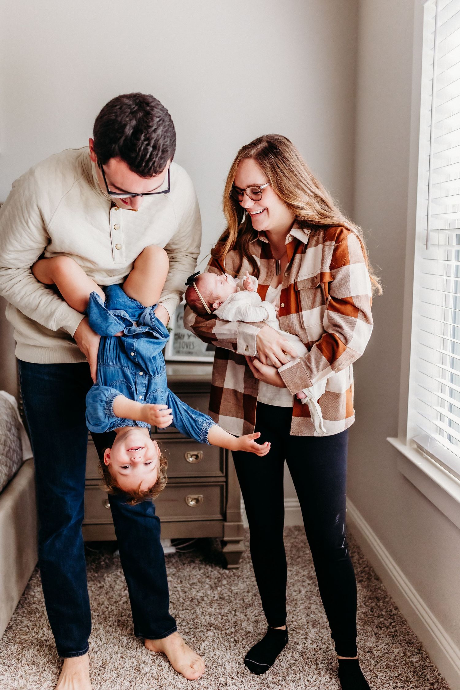 Parents playing with their newborn baby in a cozy home setting against a window.