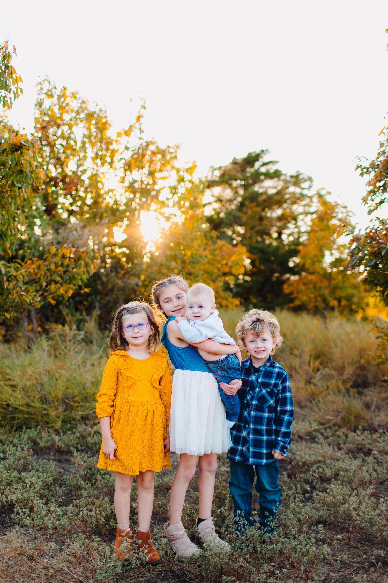 Children stand together outdoors during golden hour wearing autumn-inspired outfits in a field with fall foliage.