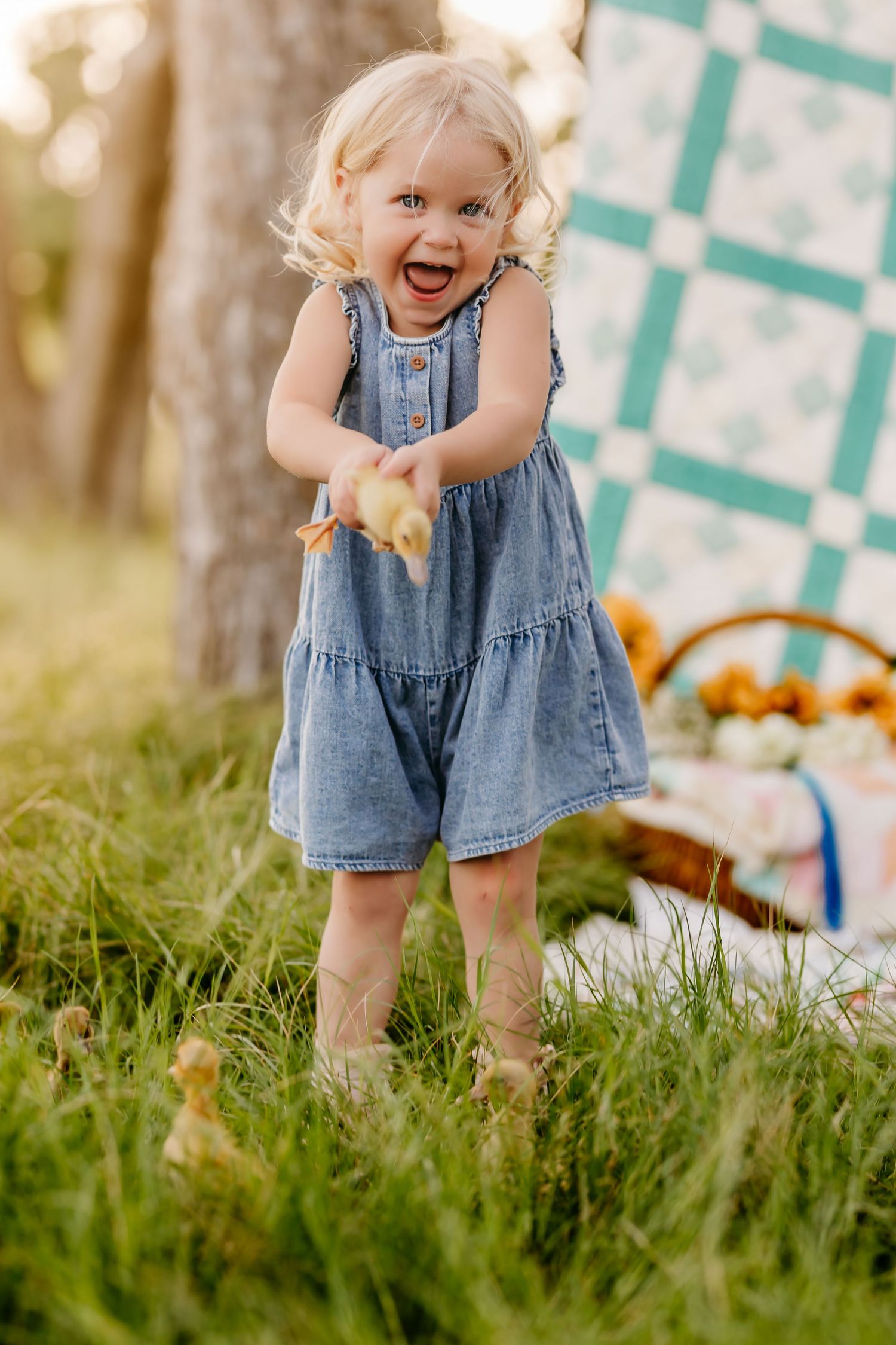 Joyful toddler girl in denim dress holding a duckling outdoors on green grass near a colorful quilt.