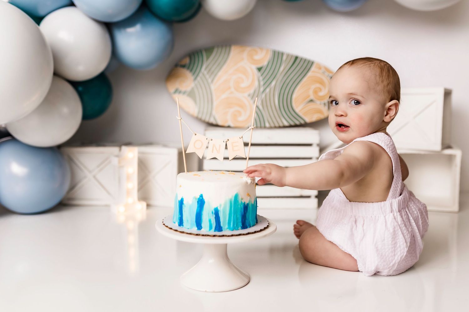 Baby in white romper reaches for blue and white birthday cake on pedestal under balloon arch decoration.