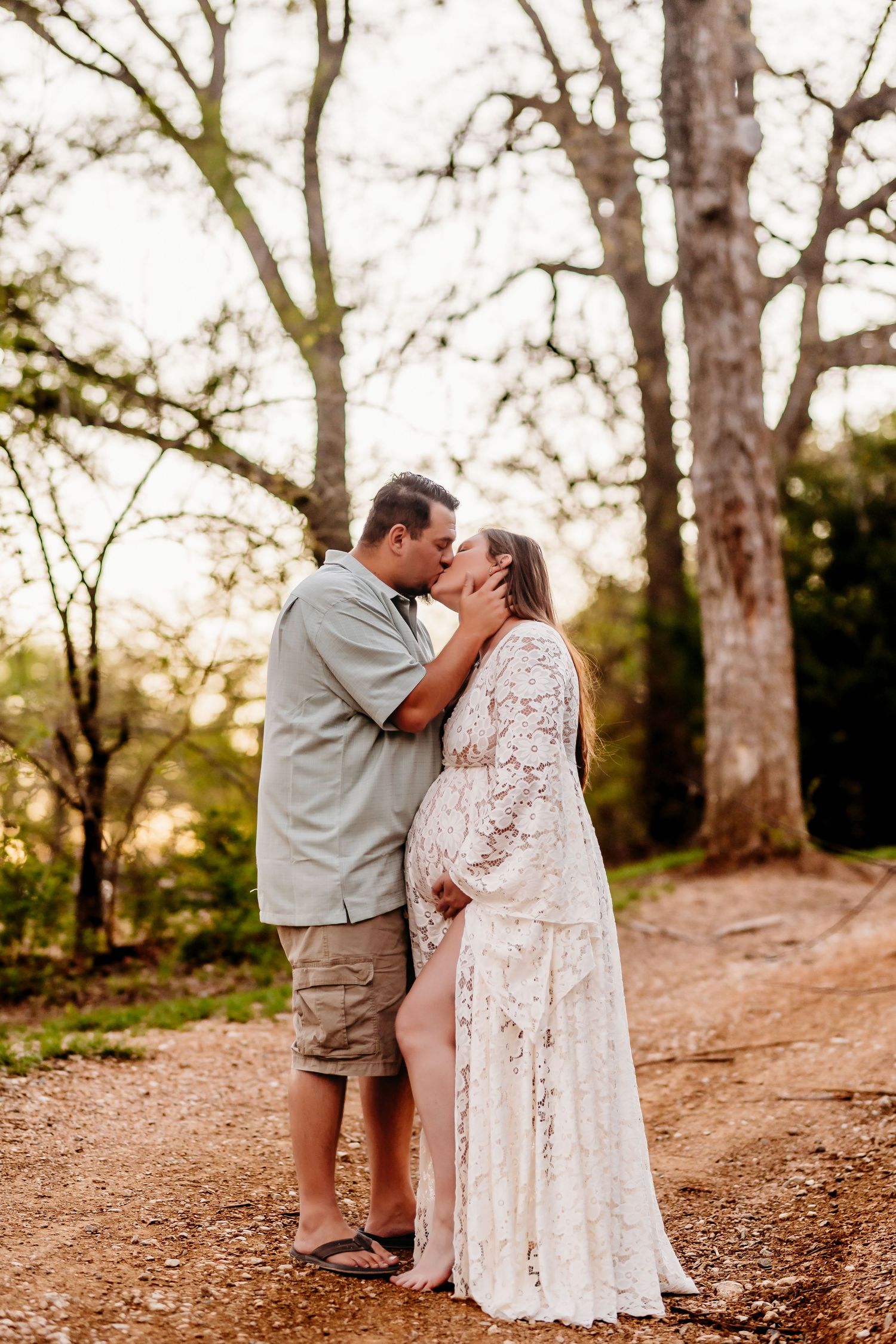 Couple sharing a tender kiss on a woodland path, woman in white lace maternity gown, man in casual shorts and shirt.
