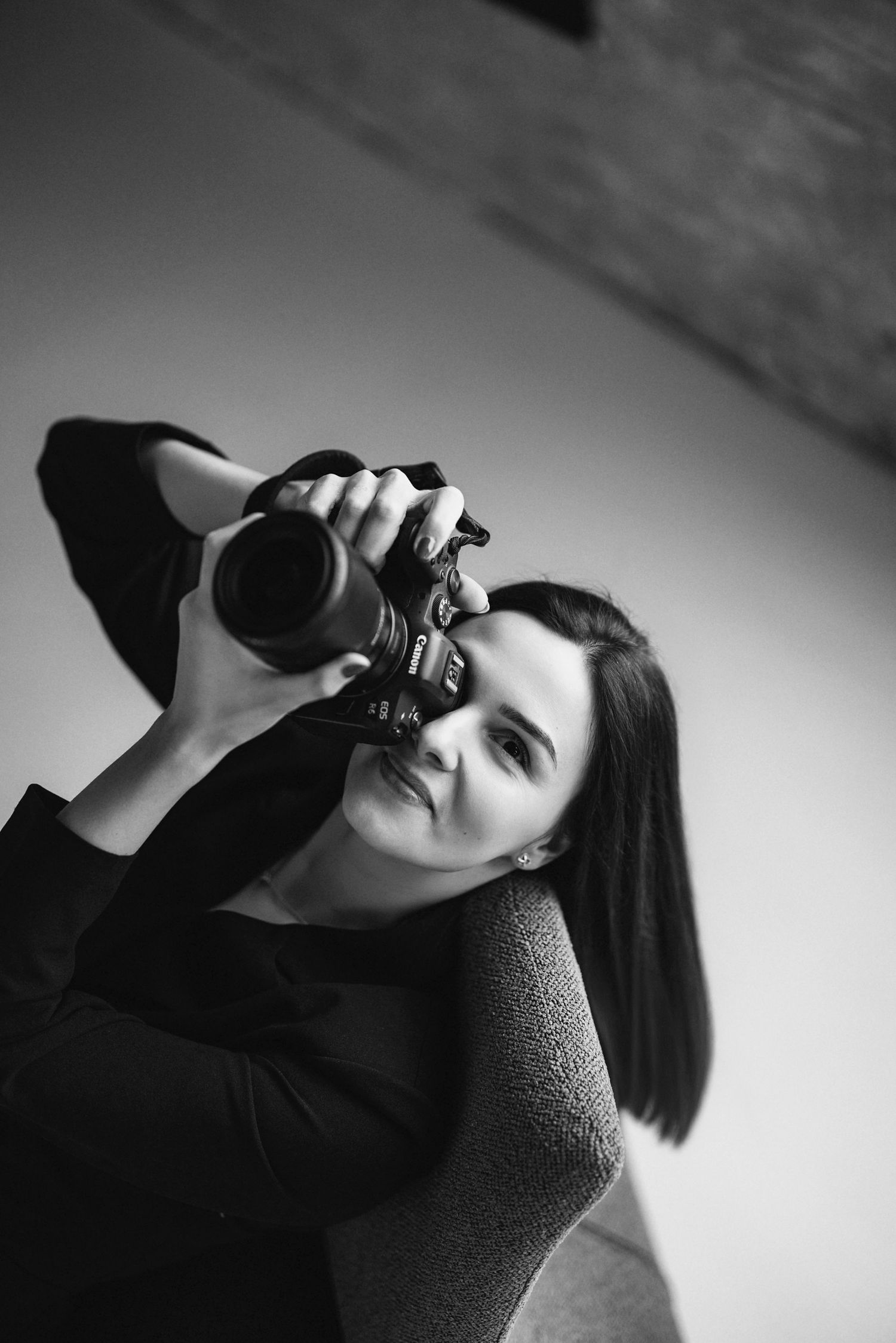Black and white photo of a woman photographer holding a DSLR camera up to her eye while seated in a chair.