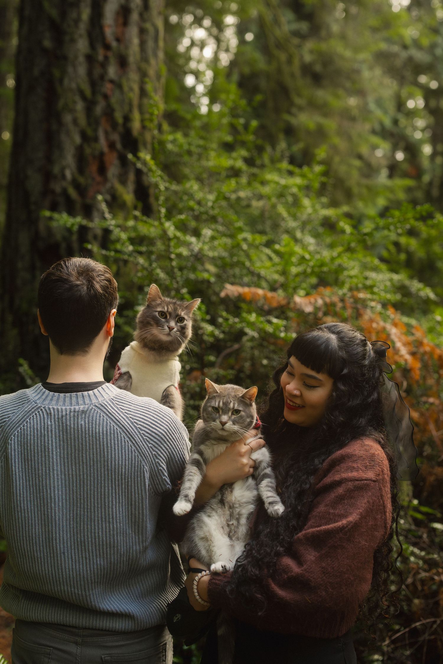 A couple in sweaters holds their gray and white cat while enjoying a peaceful moment in a forest setting.
