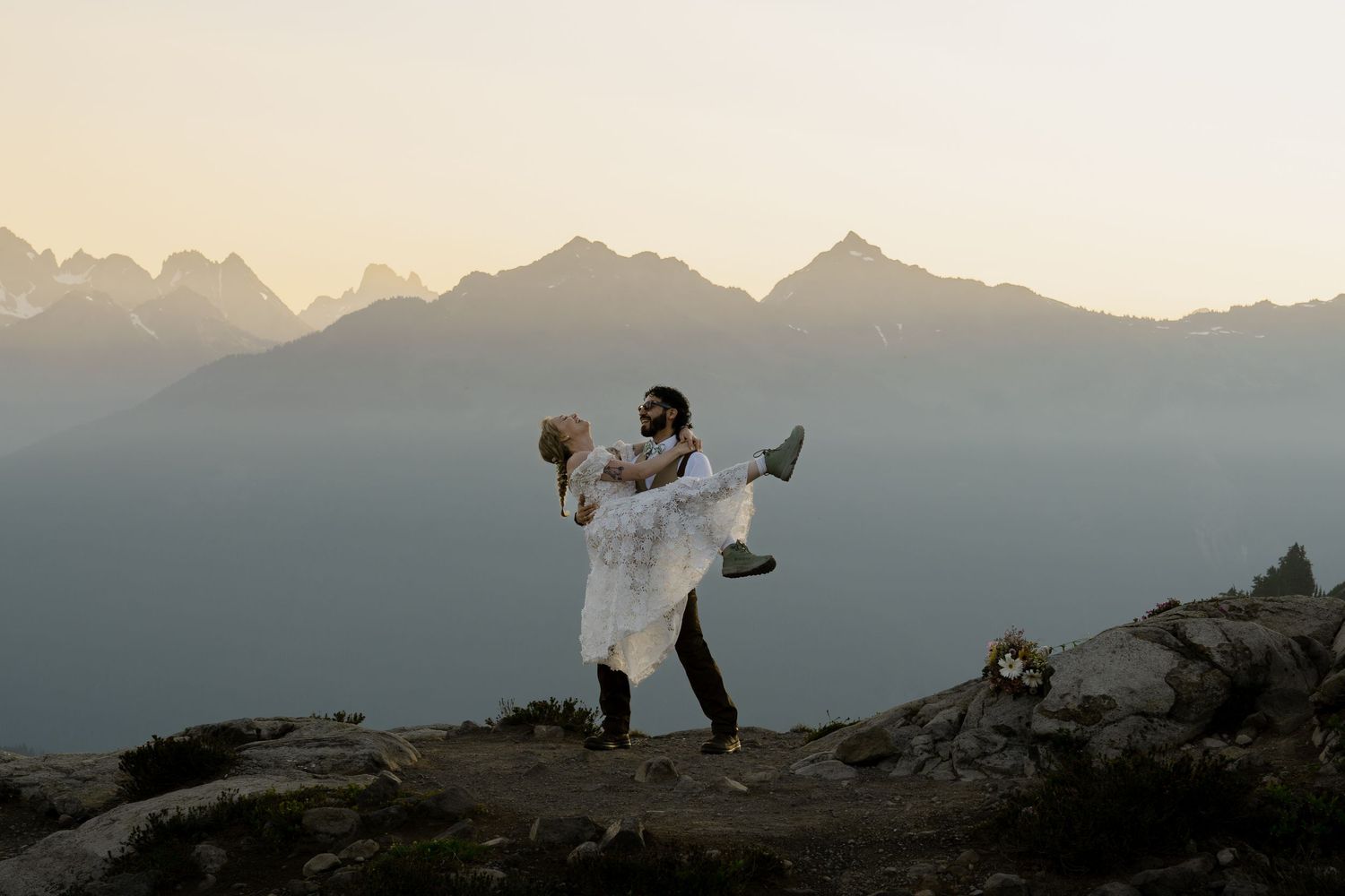 Couple celebrating wedding elopement on mountain summit with stunning alpine peaks and misty valley backdrop at sunset.