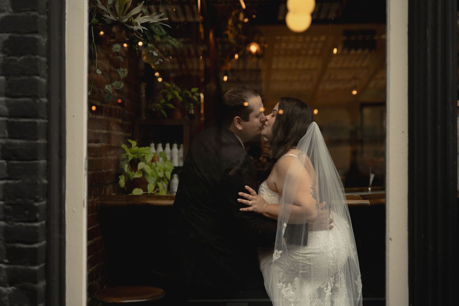 An intimate wedding kiss photographed through a window with warm ambient lighting.