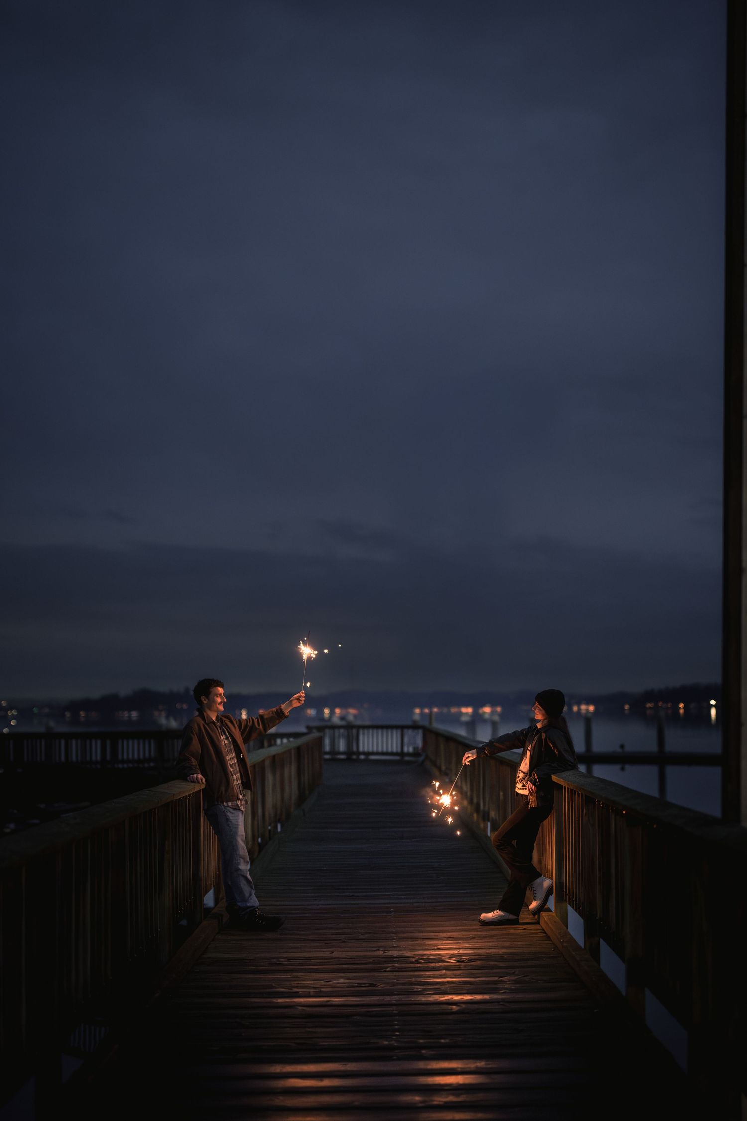 People holding sparklers create a magical nighttime scene on a wooden pier overlooking dark waters and distant lights.