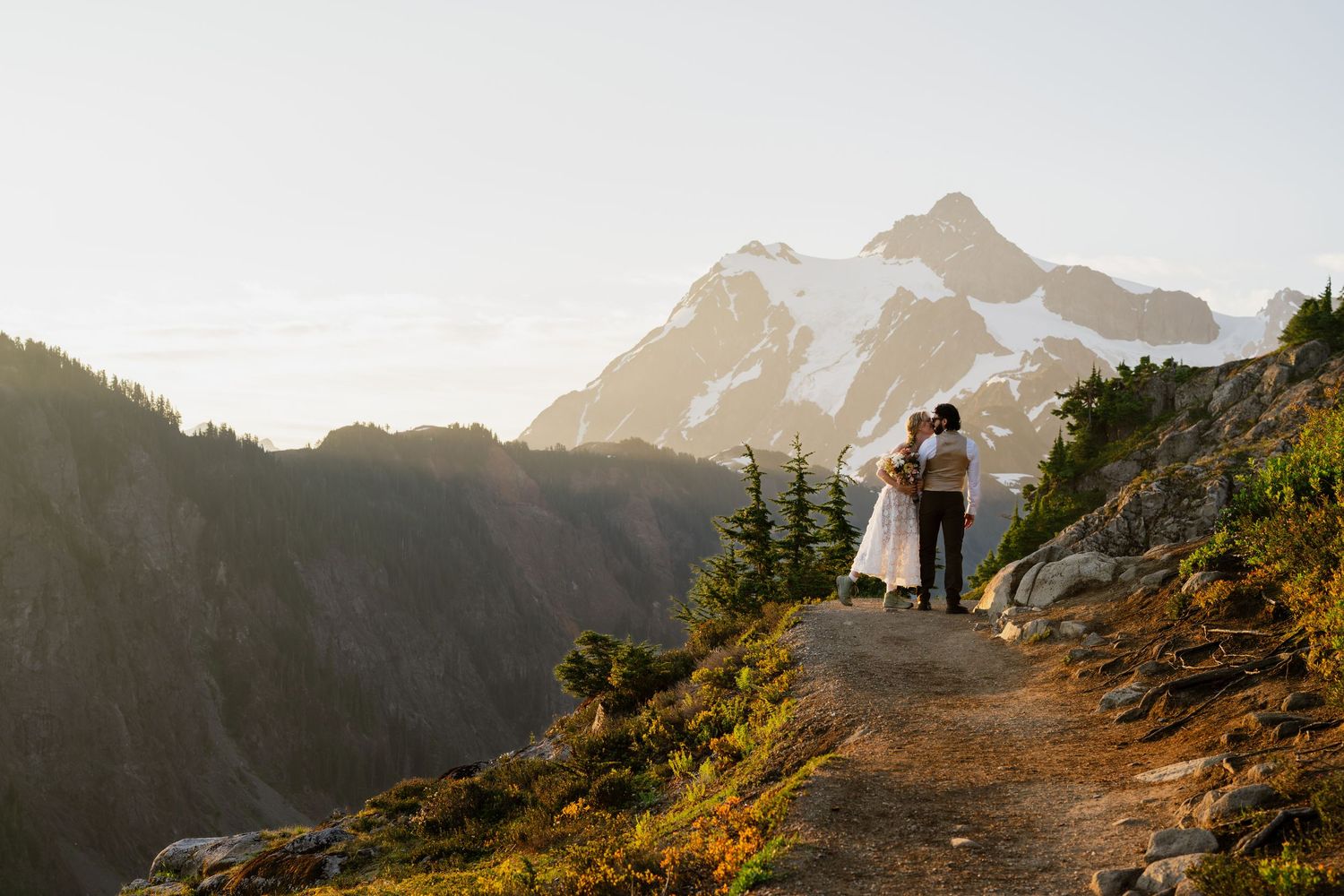 Couple embracing on a mountain trail with snow-capped peaks and golden sunset light in a scenic wilderness wedding.