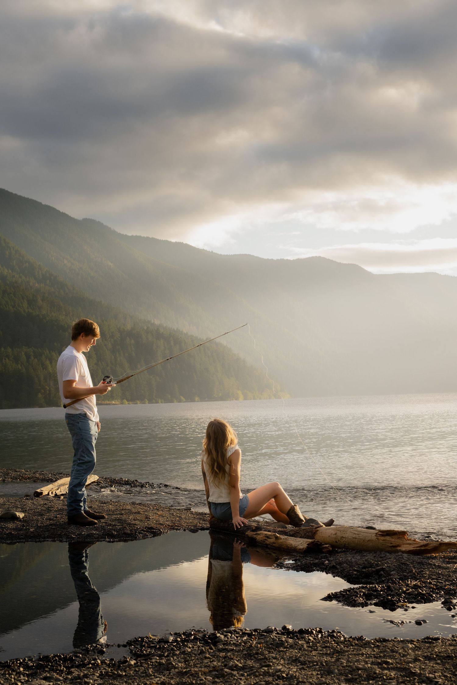 Couple fishing by a misty mountain lake at sunset, man standing with rod while woman relaxes on driftwood log.