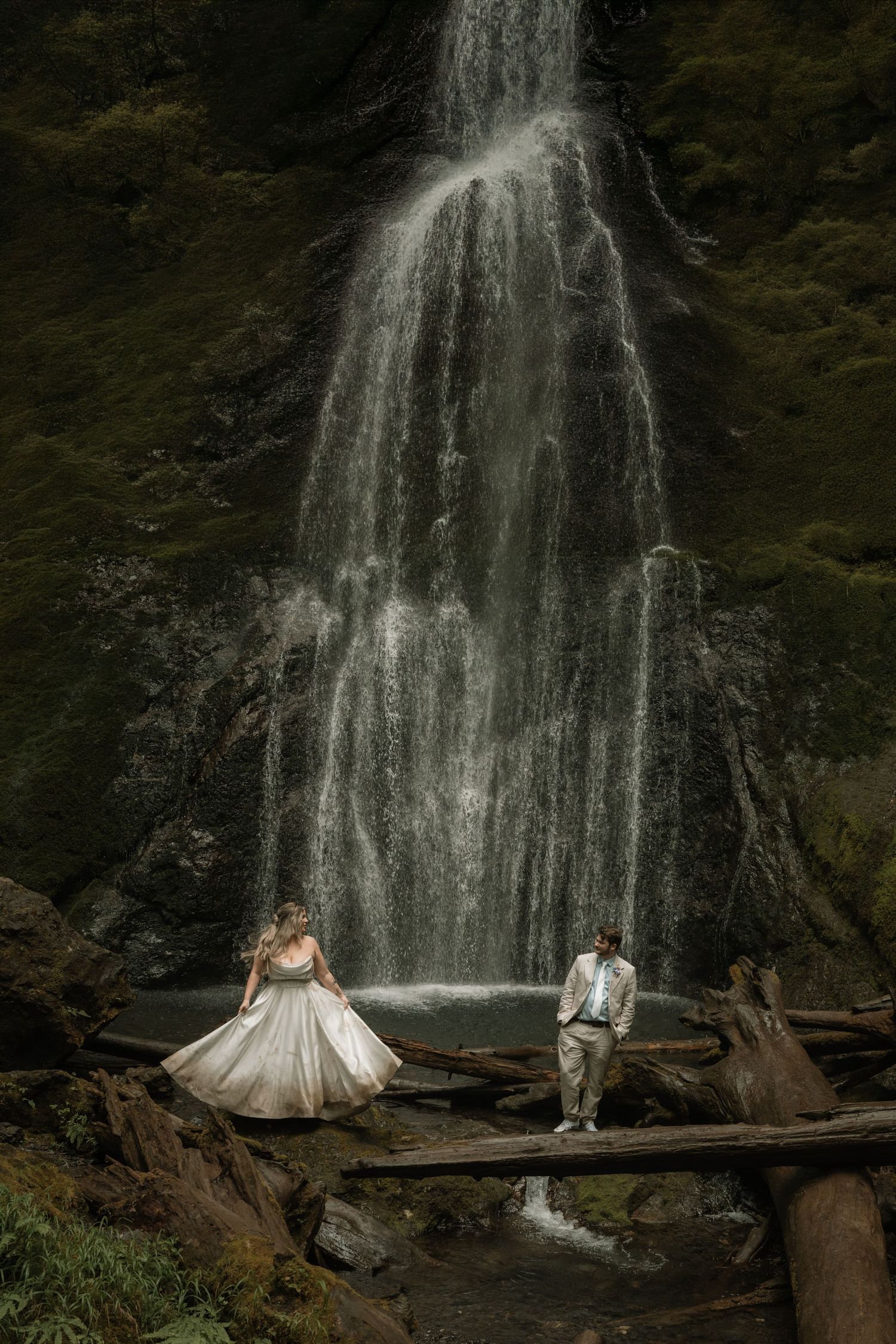 Bride and groom stand before a stunning waterfall on rustic logs in a lush forest wedding photo.