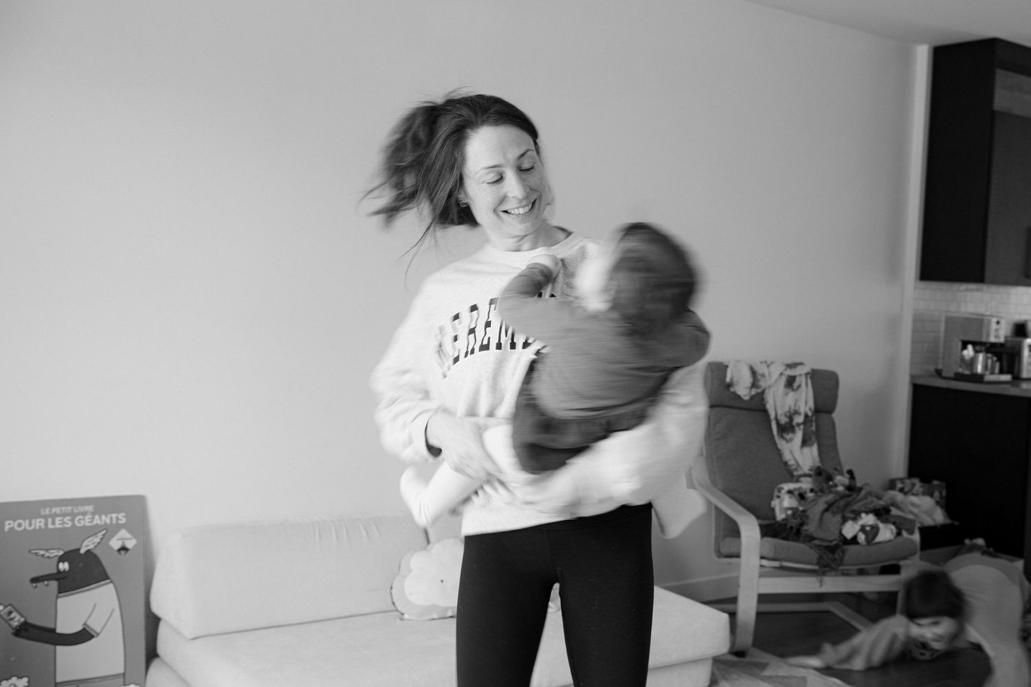Black and white photo of a laughing woman playfully lifting a child in a cozy living room.