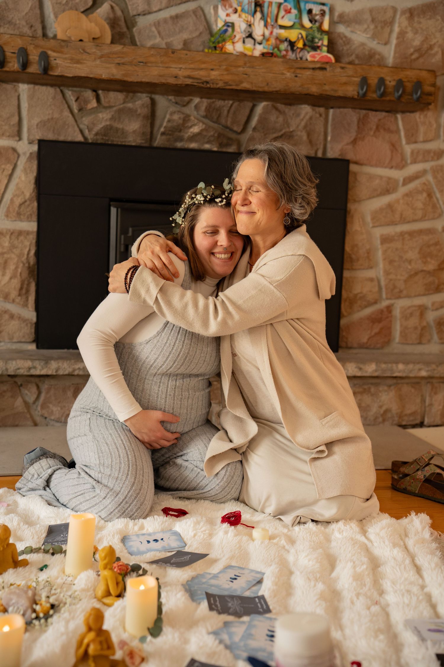 Pregnant woman and older woman hugging warmly by a stone fireplace, surrounded by candles and keepsakes.