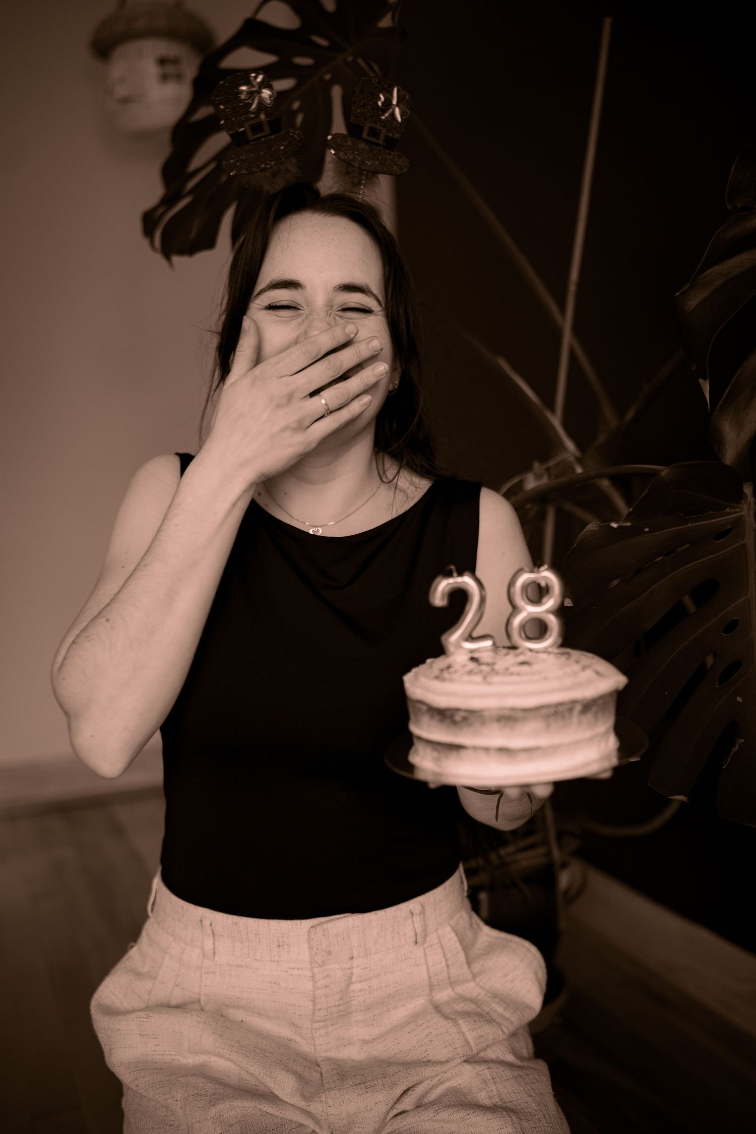 Young woman laughing and holding a birthday cake with number 28 candles, wearing a crown in sepia-toned photo.