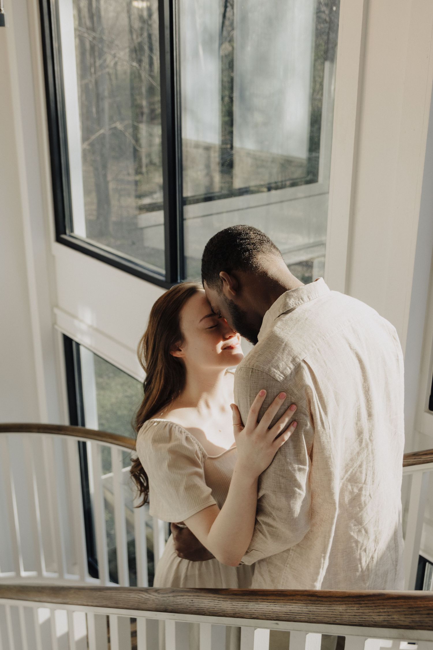 A romantic couple embraces near a sunlit window in matching cream colored outfits.