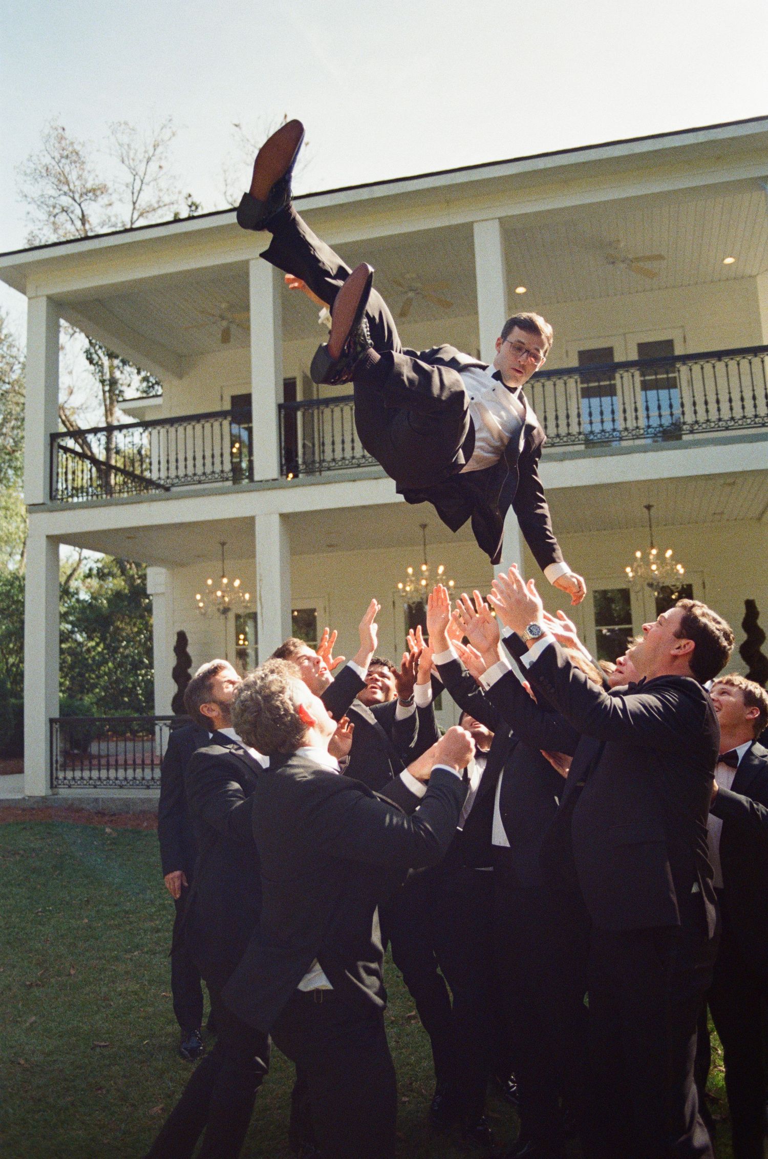 Groomsmen tossing groom into the air in front of elegant two-story white plantation venue during wedding celebration.