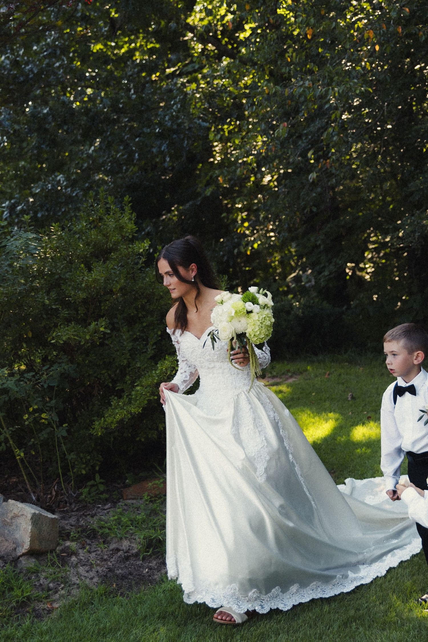 A romantic outdoor wedding scene with a bride in a flowing white gown kneeling beside a young ring bearer in nature.