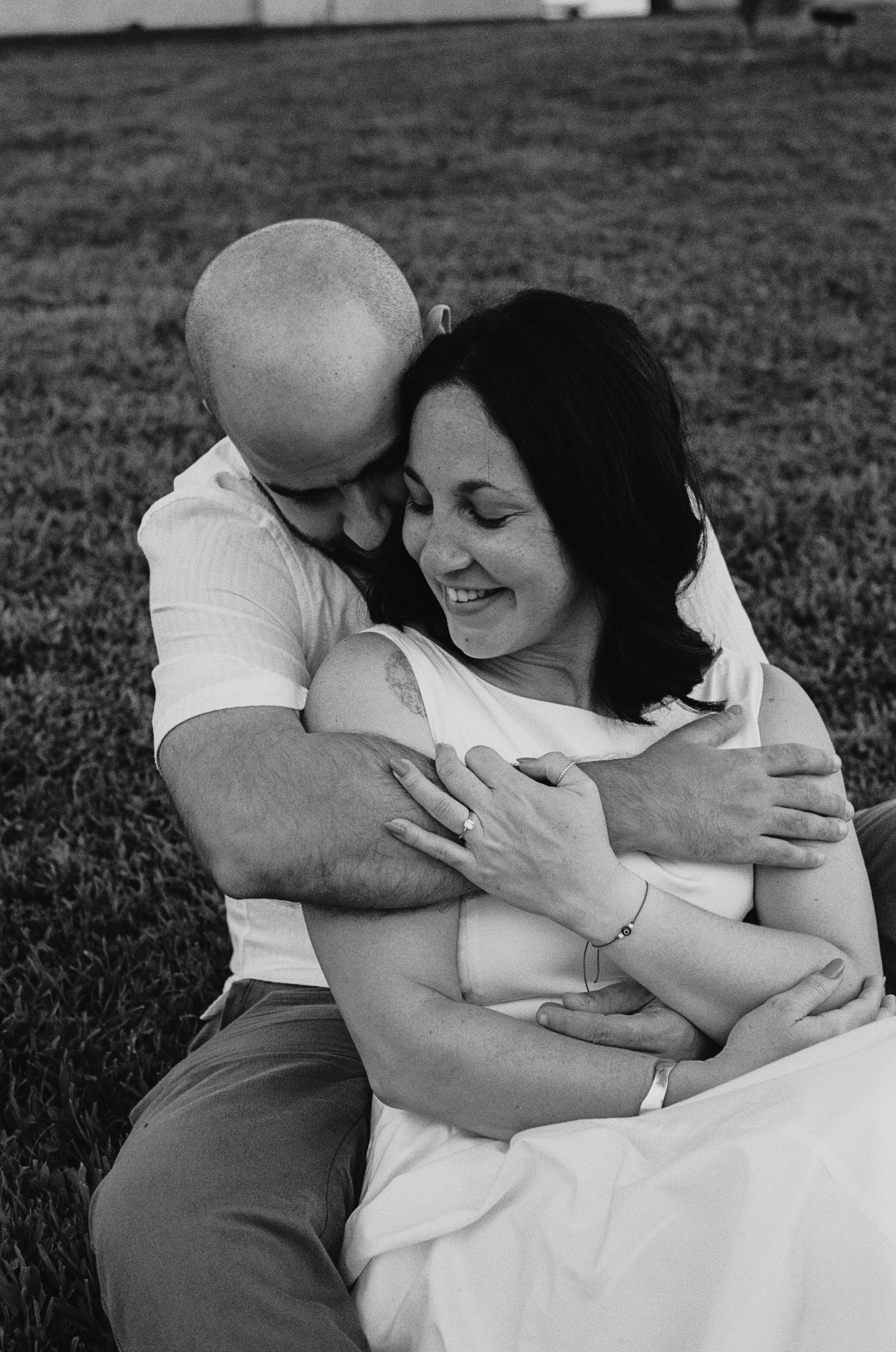 Black and white photo of a couple embracing outdoors, man hugging woman from behind as she smiles joyfully.
