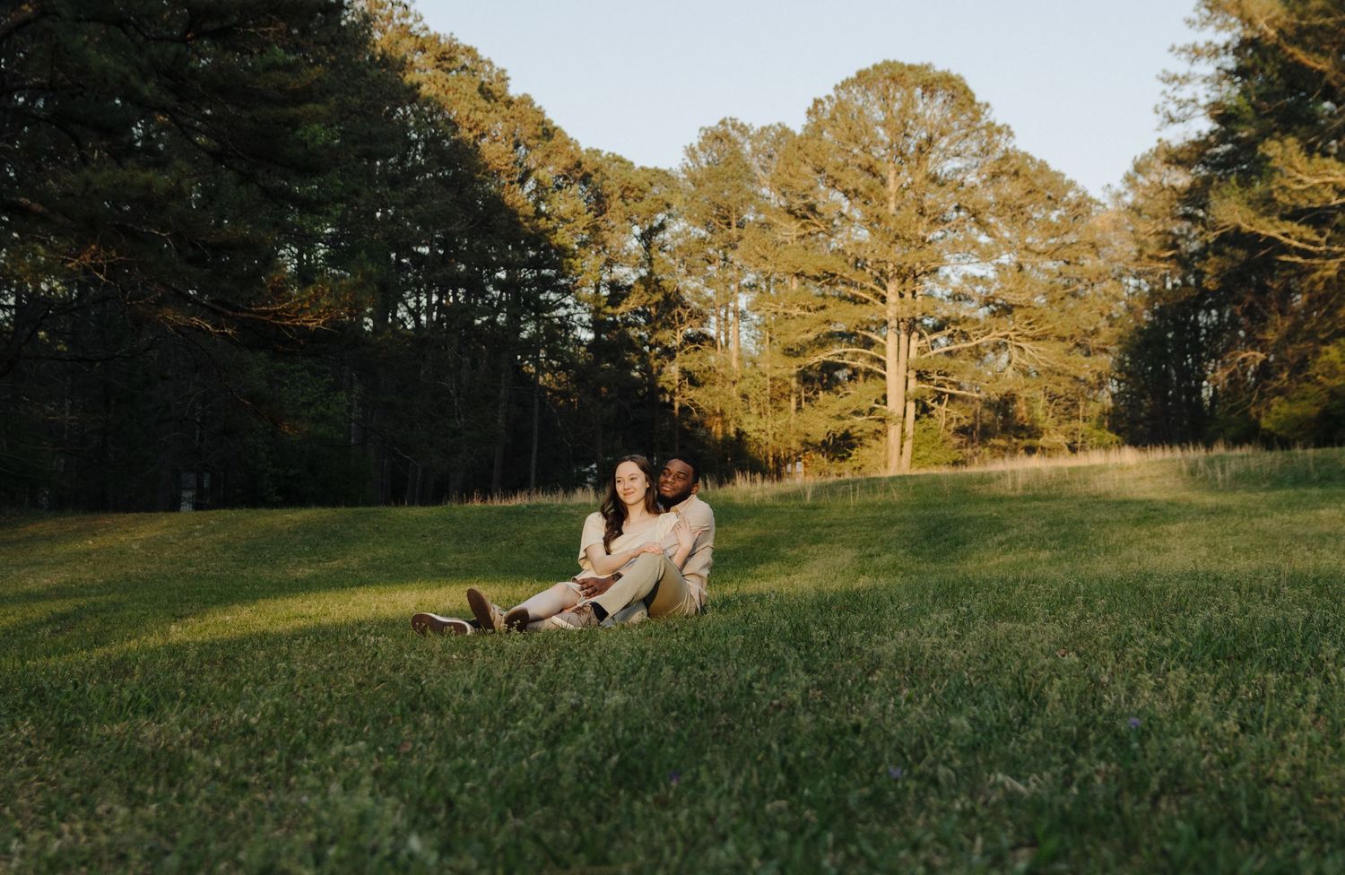 A figure relaxes on a grassy slope during golden hour with tall pine trees creating a serene backdrop.