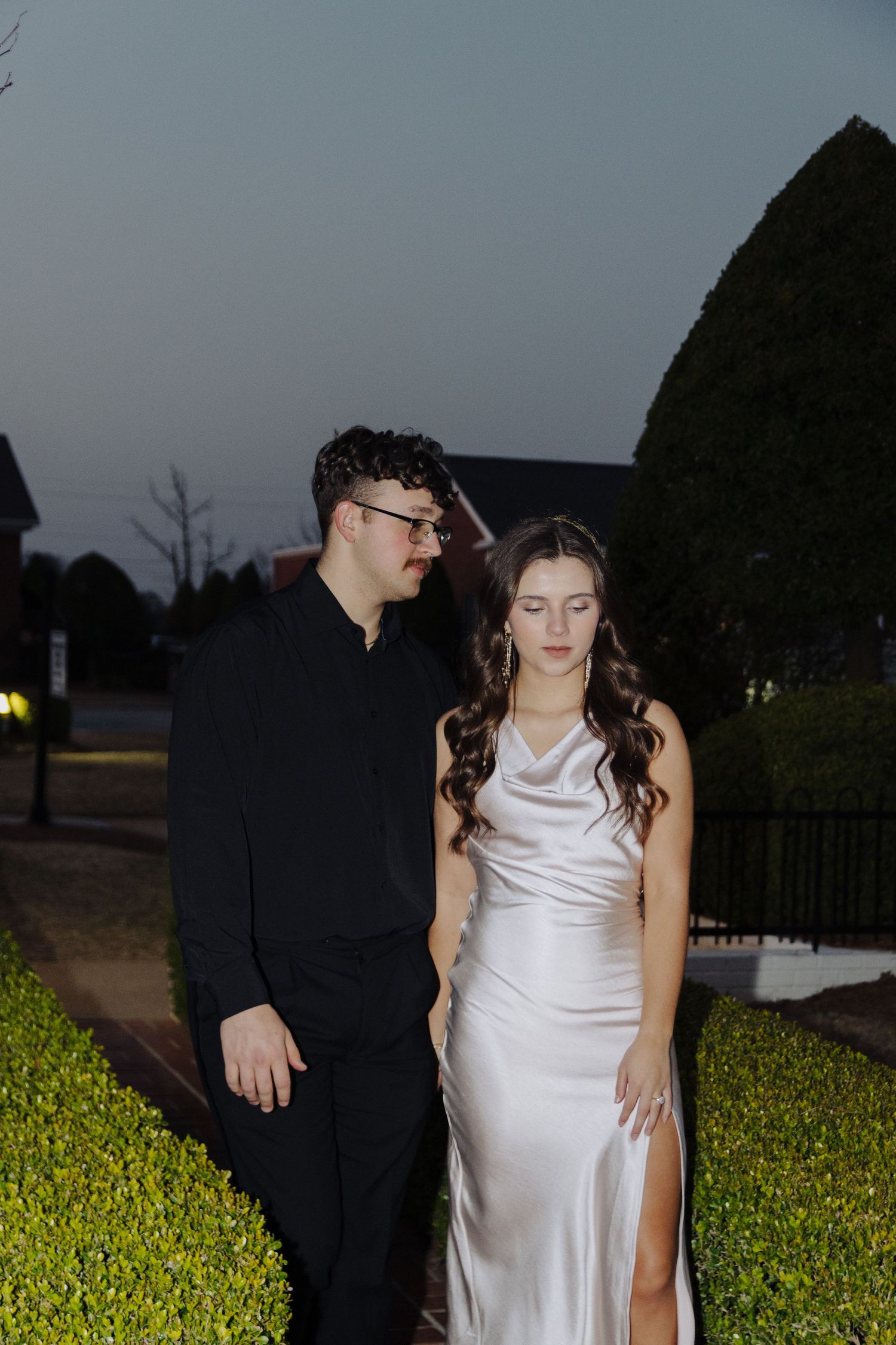A couple in formal attire poses outdoors at night against a hedge for a romantic photoshoot.