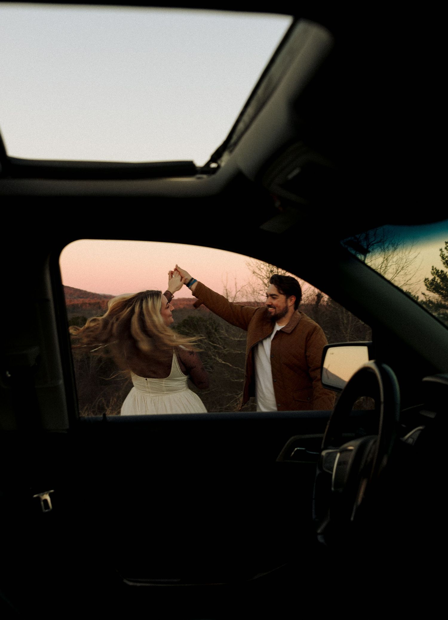View through car window of two people dancing together at sunset on a rural road.