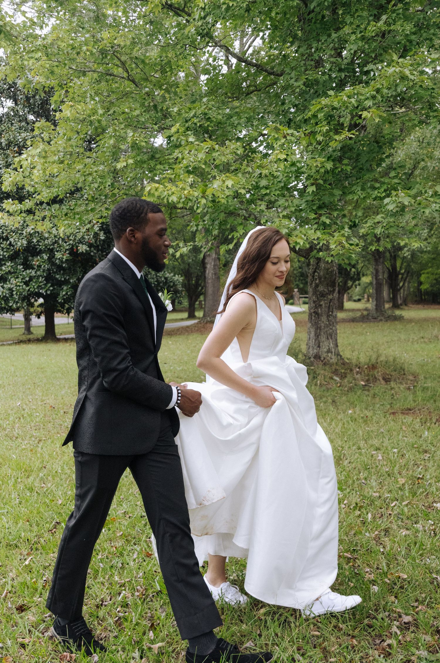 A couple in formal wedding attire walking together on grass with green trees in the background.