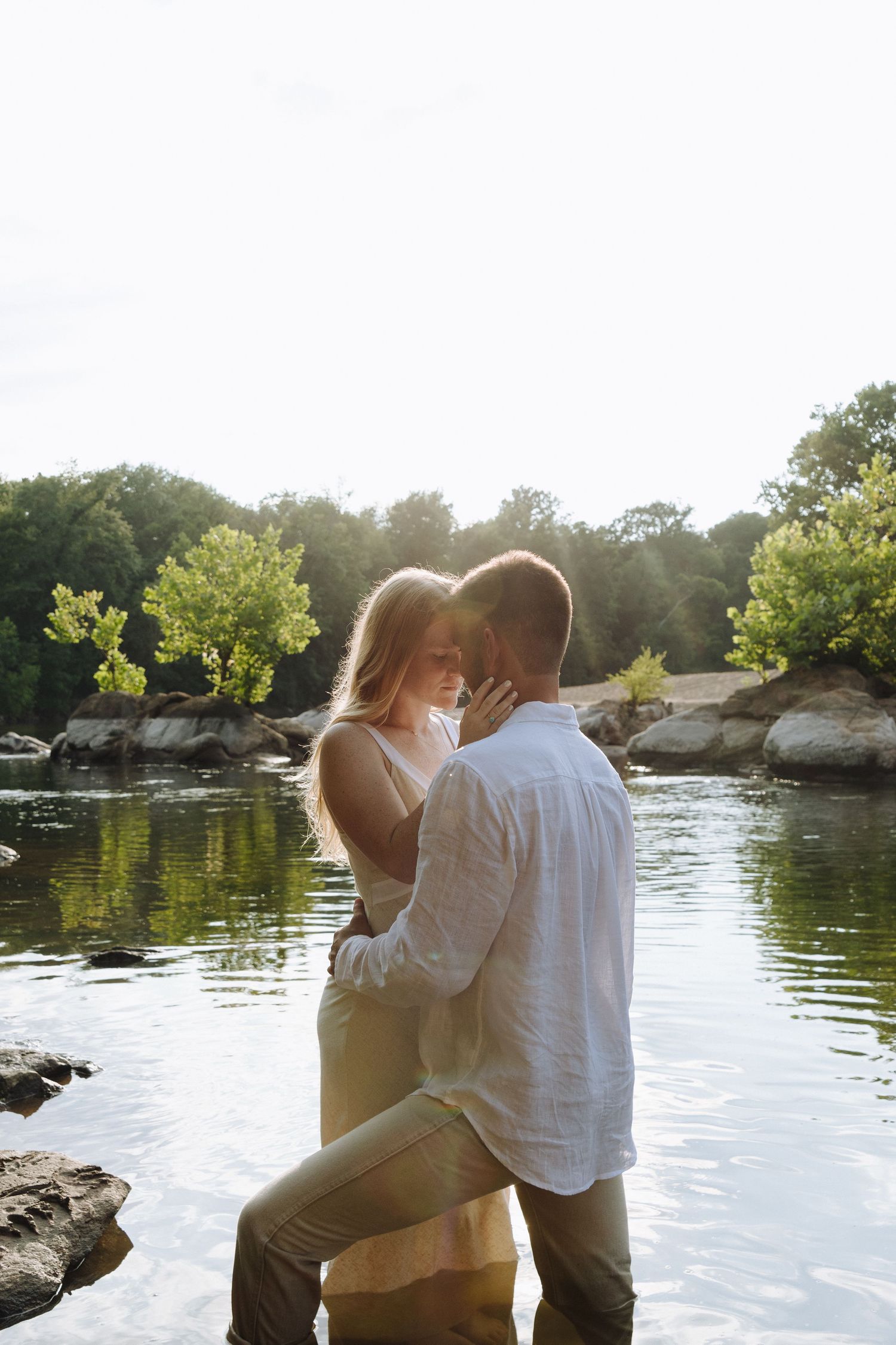 A couple sits together by a serene lake during golden hour as sunlight sparkles on the rippling water.