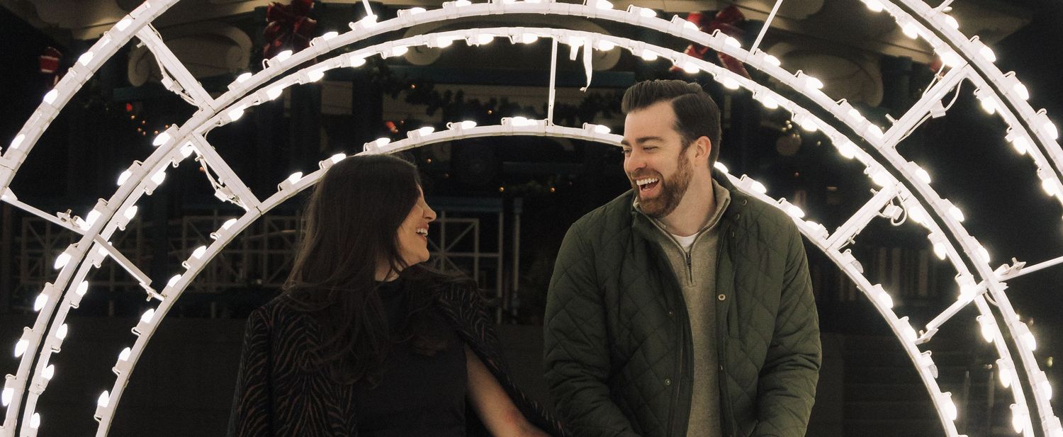 Two people laugh together while standing under illuminated round archways decorated with holiday string lights.