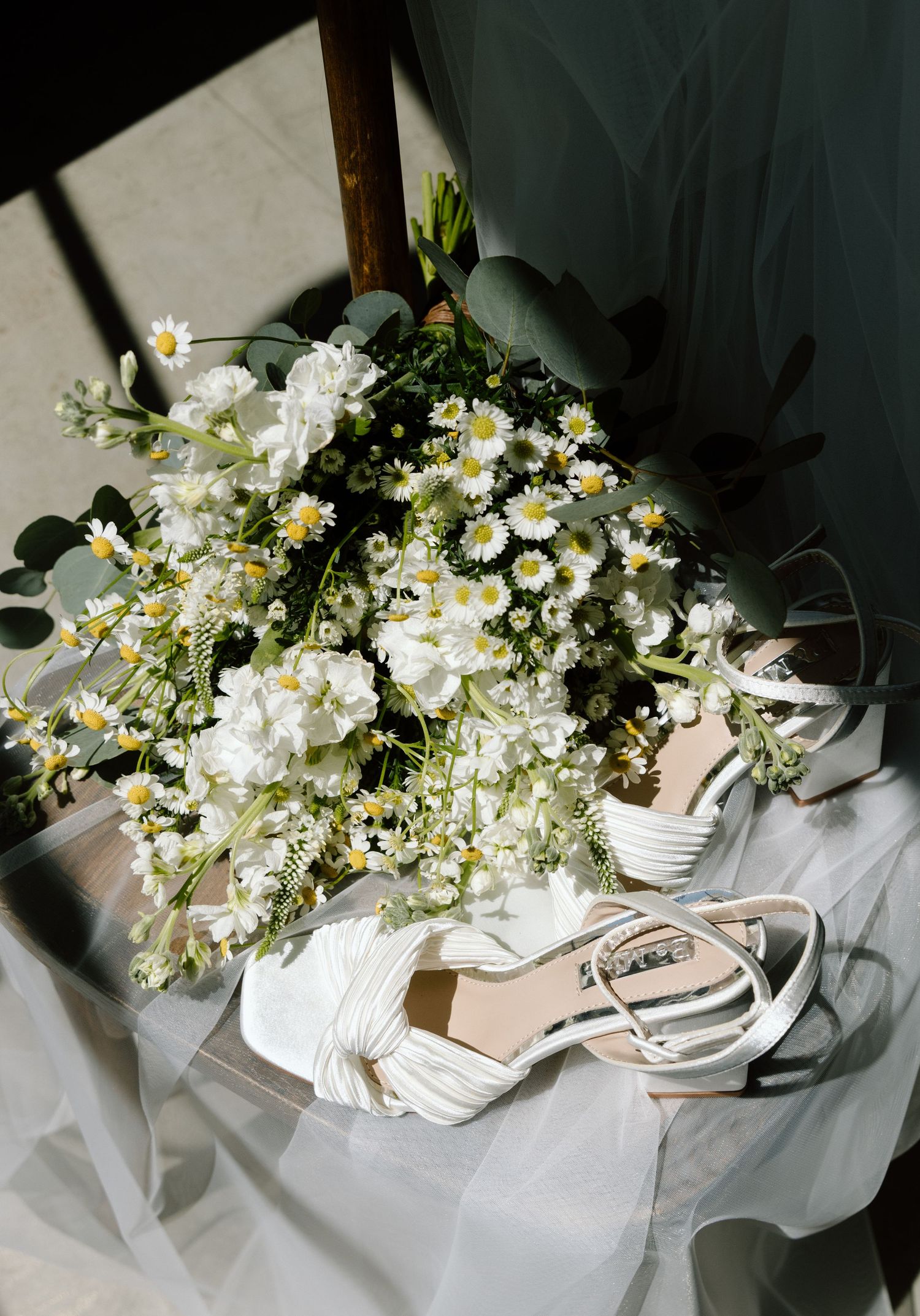 White bridal bouquet with daisies and flowers beside strappy heeled wedding shoes on a chair with tulle.