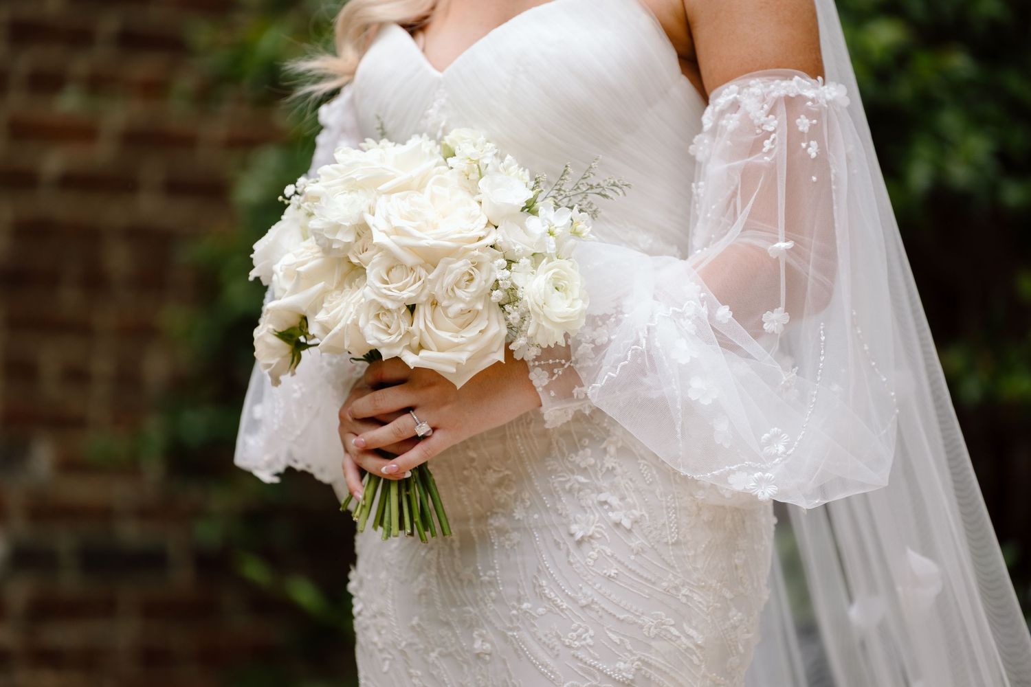 A bride in a white lace wedding dress holds a bouquet of white flowers while standing near a brick wall.