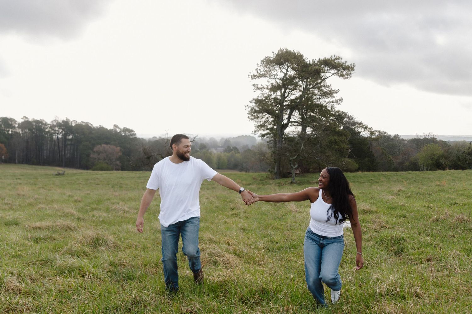 Two people walk through a grassy field holding hands on an overcast day with a lone tree in the background.