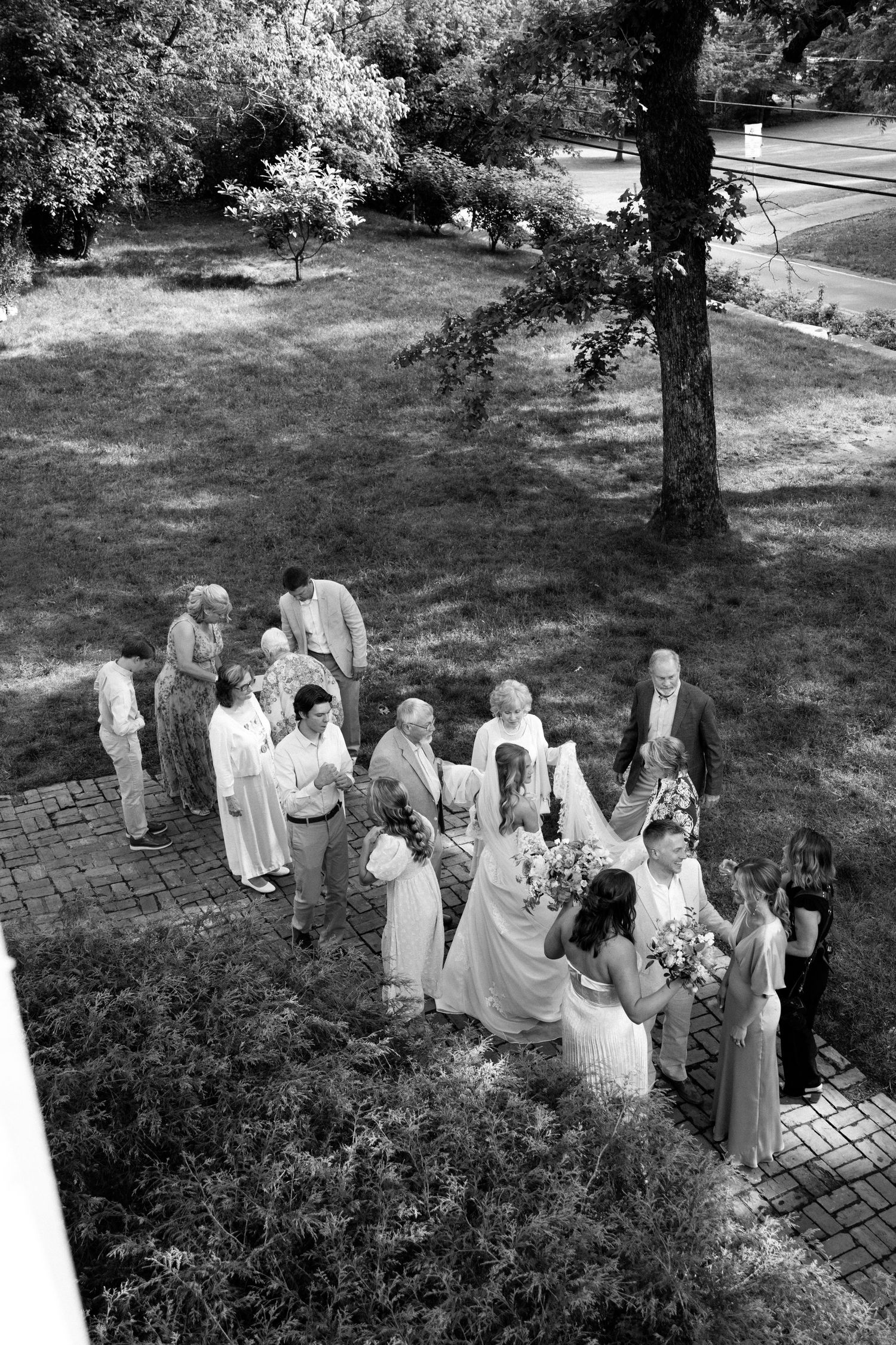 Black and white aerial view of a wedding party gathered on brick path surrounded by lush greenery.