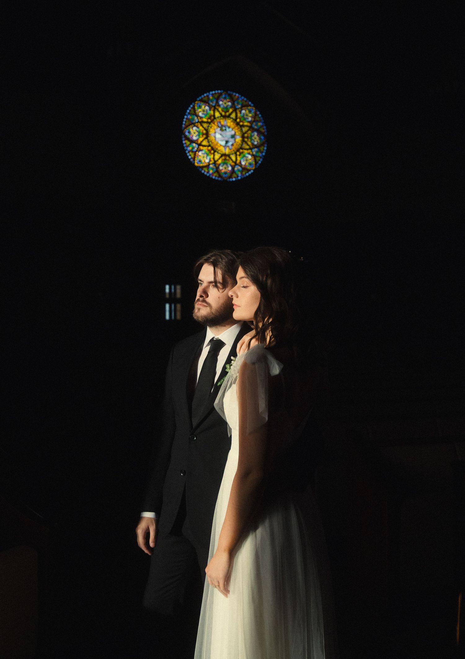 A romantic wedding couple shares an intimate moment under a stained glass window in a dark, dramatic setting.