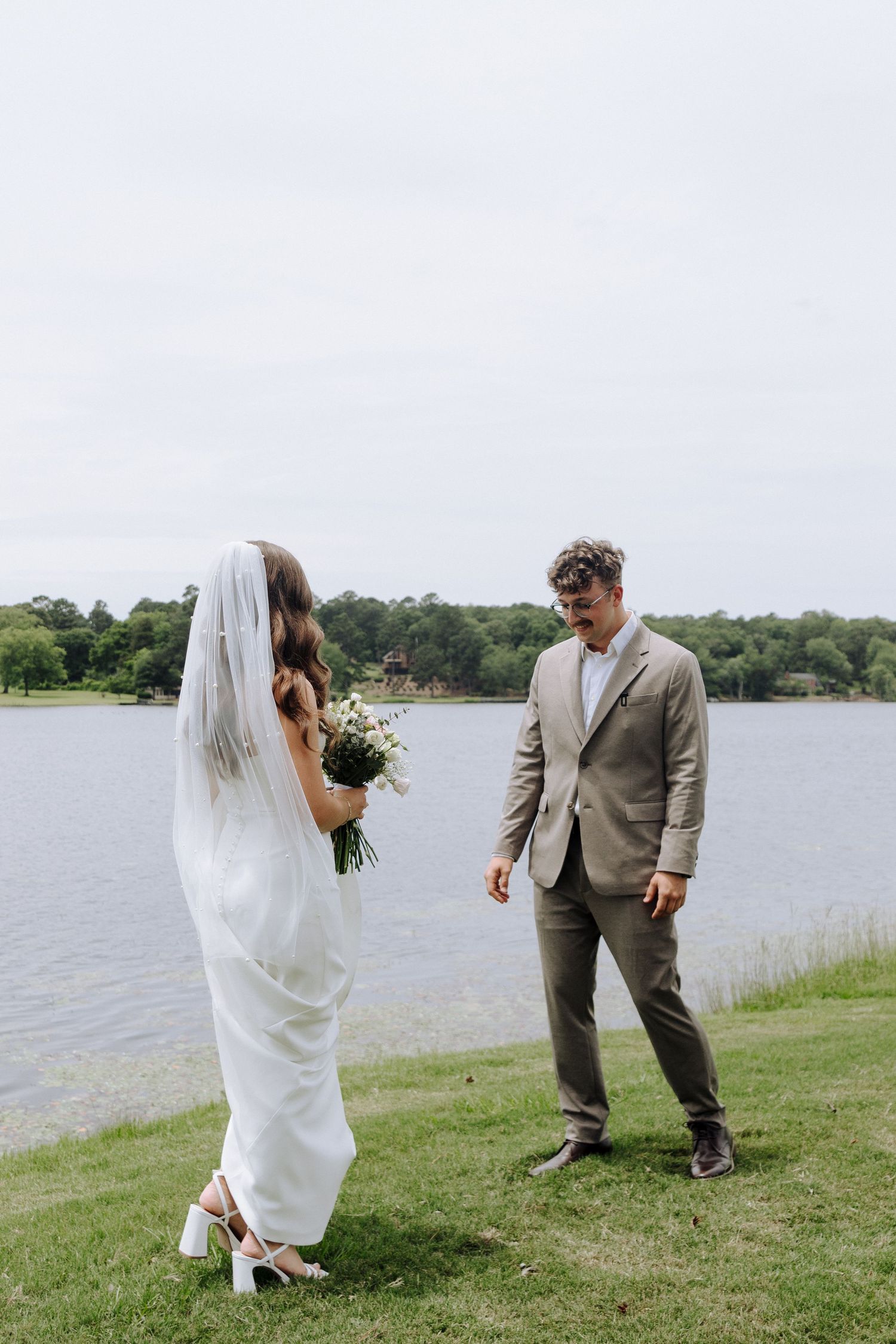 A couple shares a candid wedding moment by a serene lakeside, with the bride in white and groom in beige.