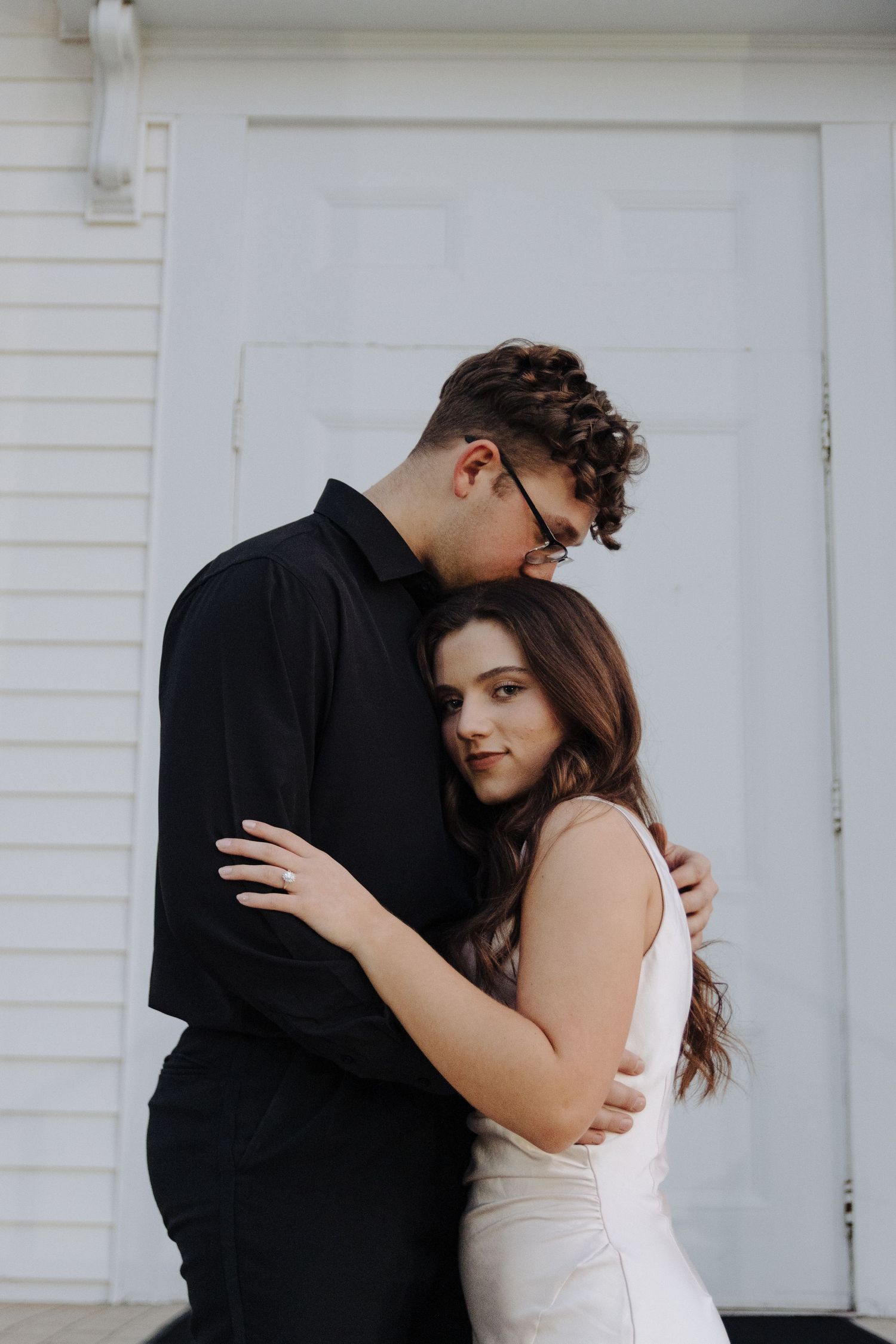 A romantic couple embraces against a white door, with one wearing a black shirt and the other in white.