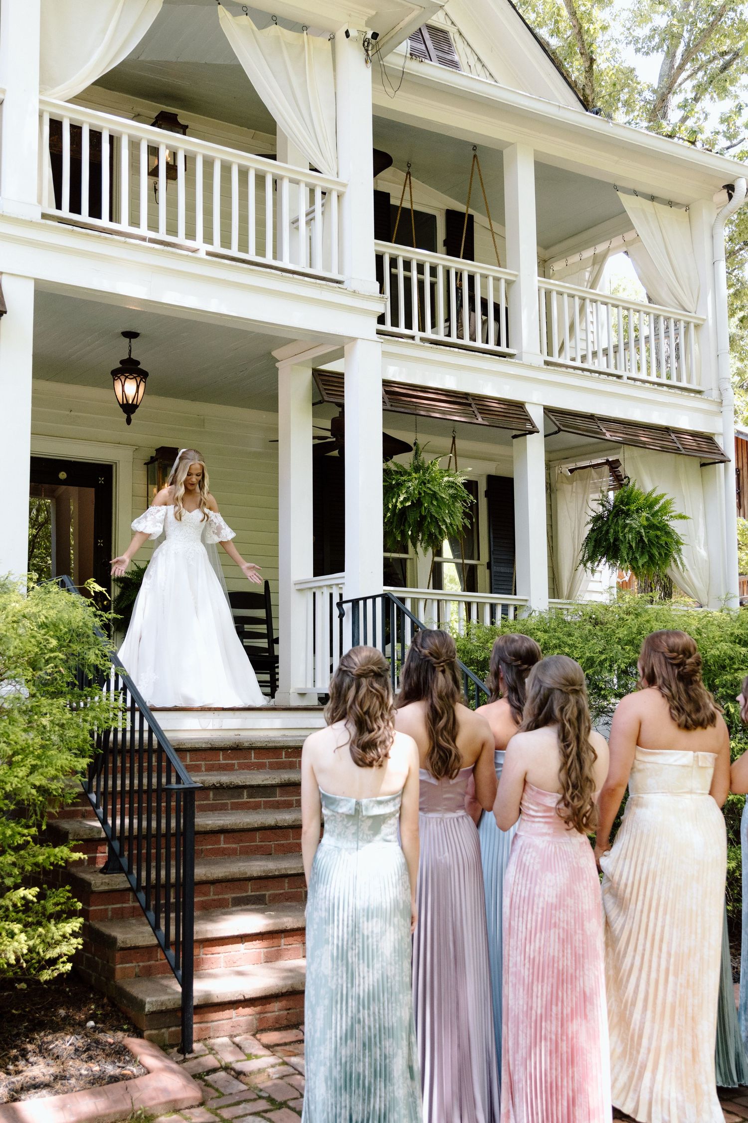 Bride in white gown stands on brick steps of white colonial home as bridesmaids in pastel dresses look on.
