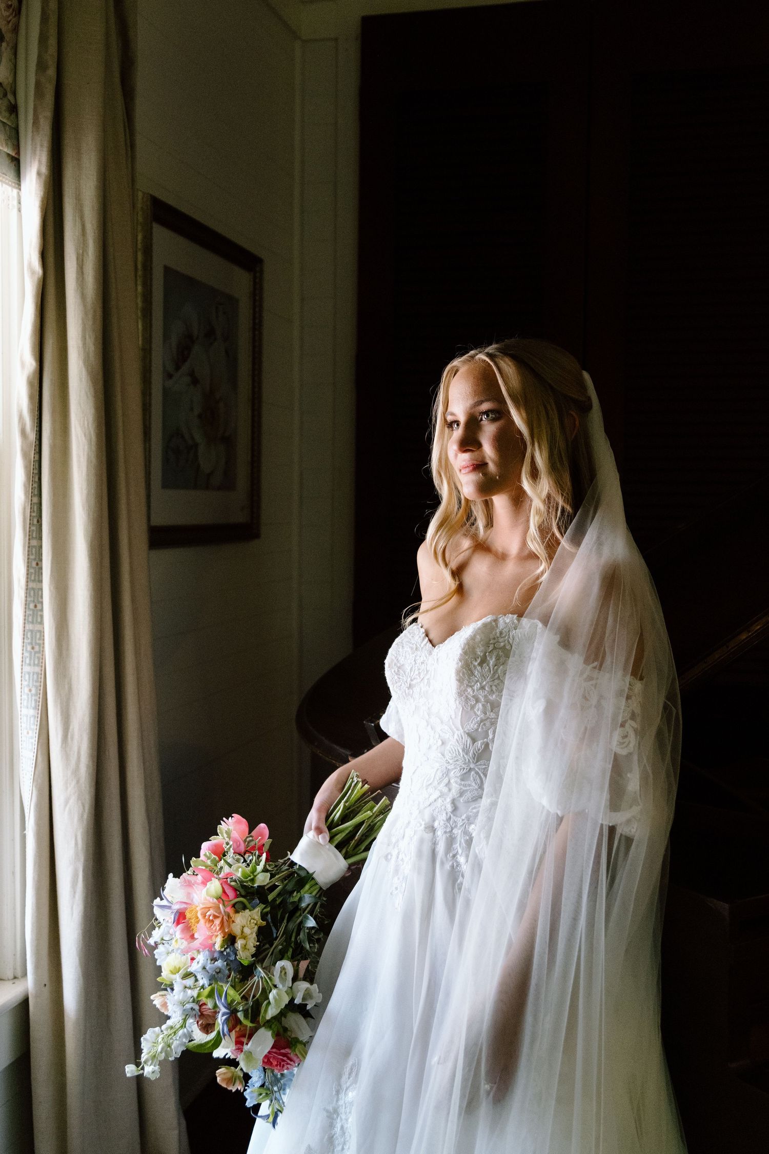 Bride in white lace gown and veil holds colorful bouquet by window in elegant dark interior.