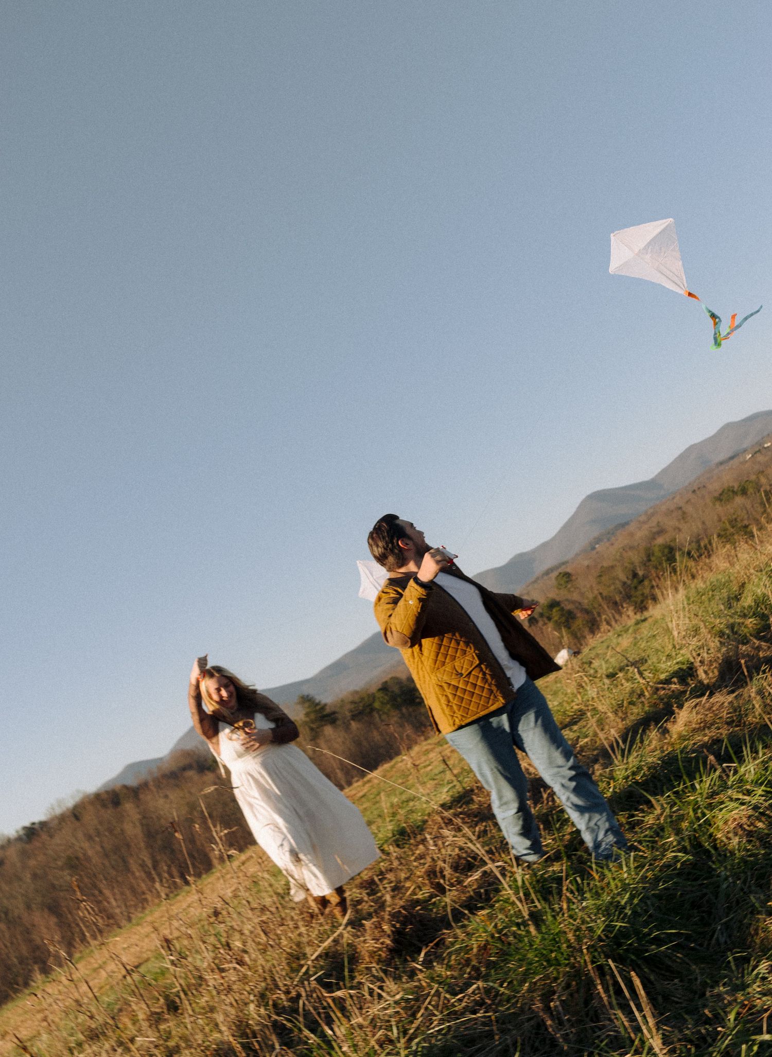 Two people fly a white kite together on a grassy hillside against a mountain backdrop on a sunny day.