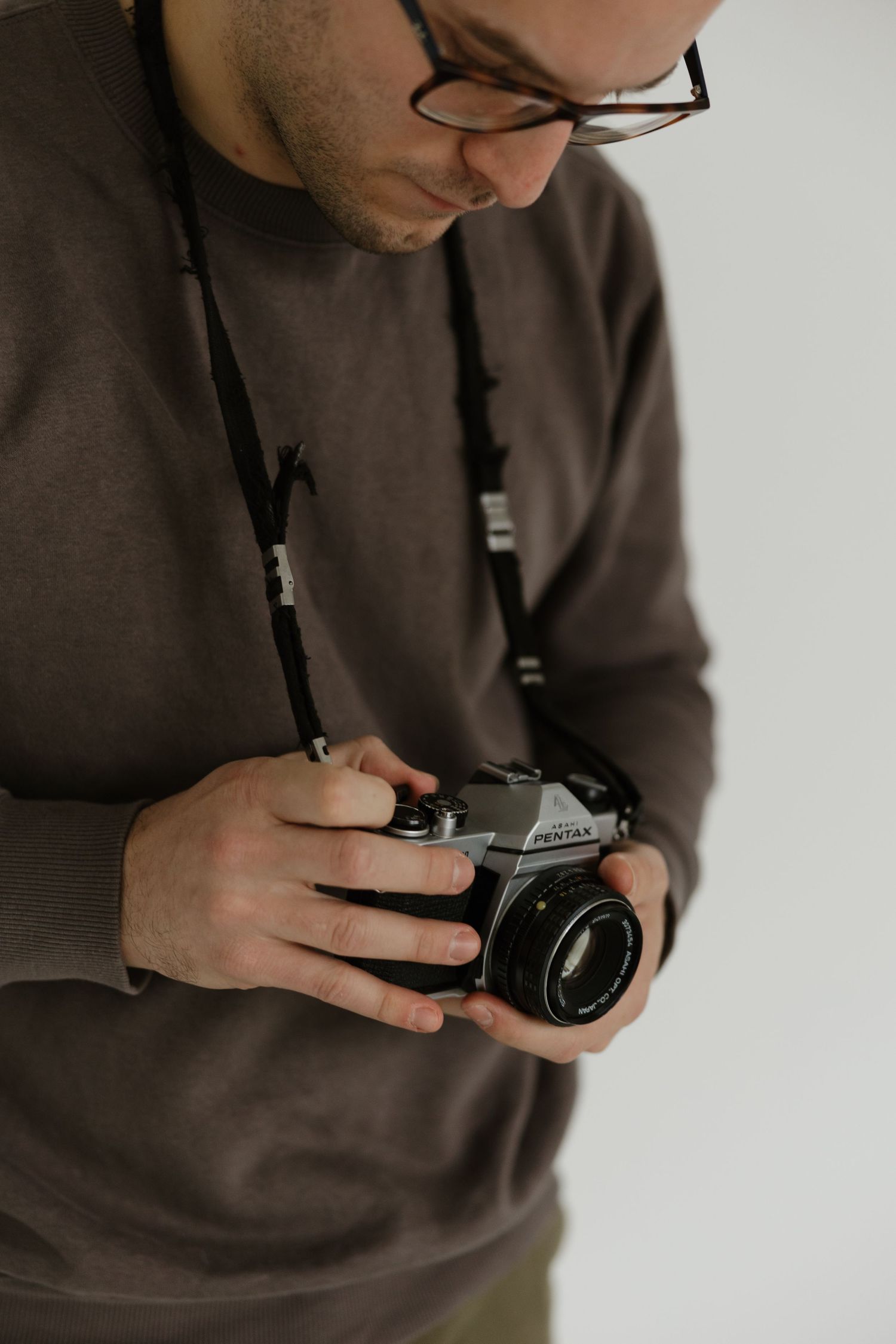 A person wearing glasses and a brown sweater holds a vintage camera against a light background.
