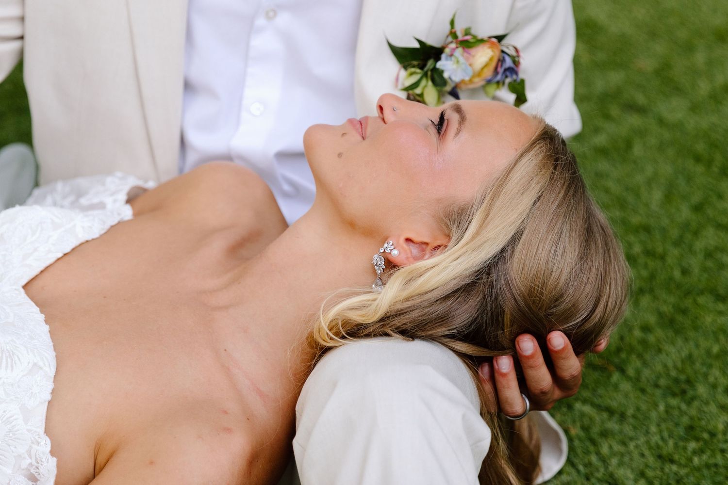 Bride with blonde hair lying on grass, wearing lace dress and earrings, groom in white suit gently holding her head.