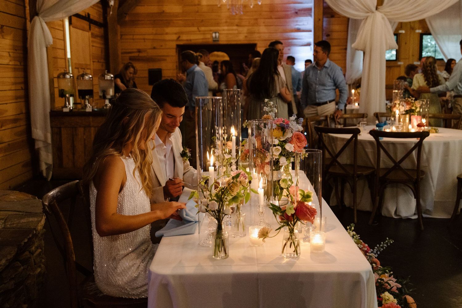 Couple at candlelit wedding reception table with floral centerpieces in a rustic barn venue.