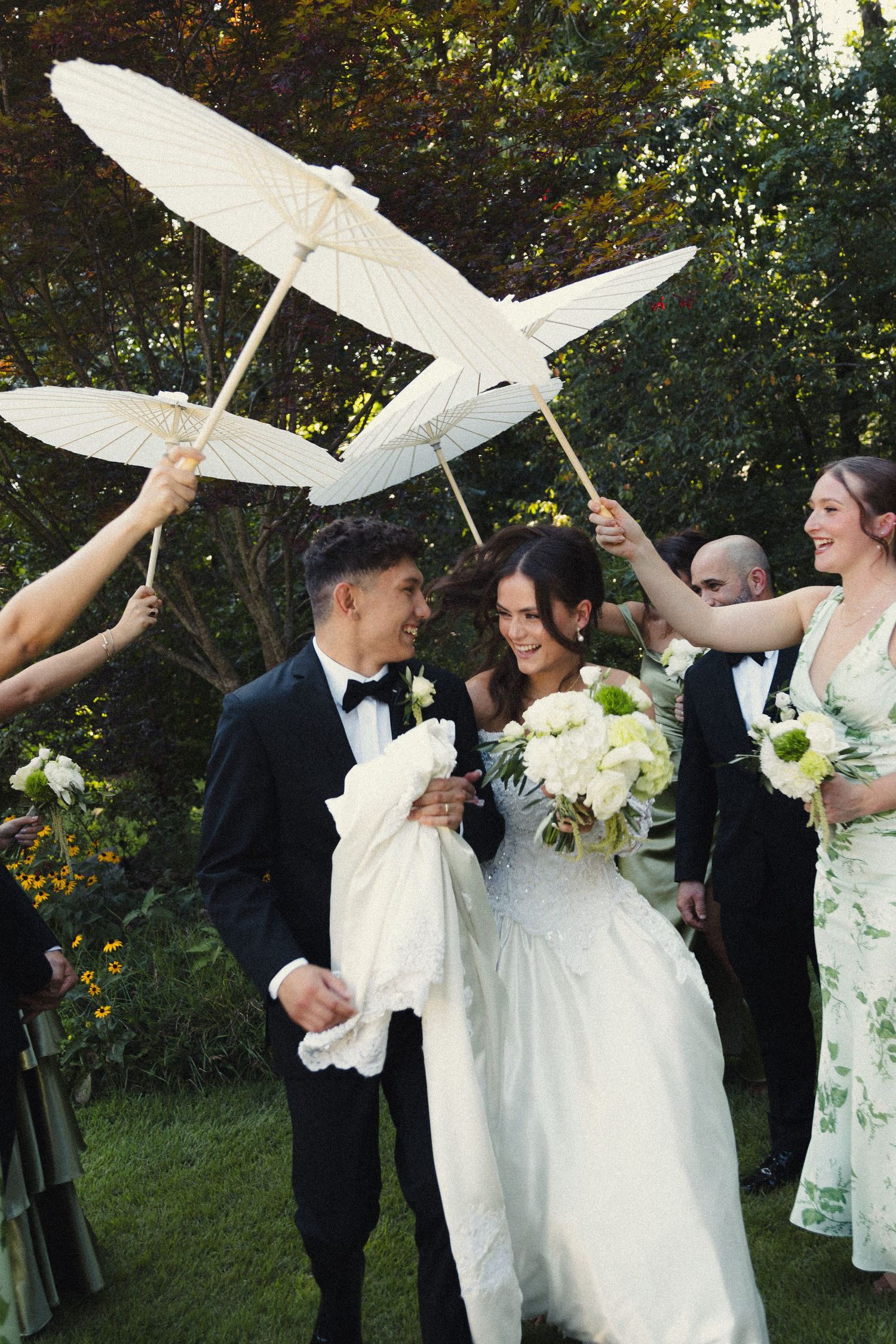 Wedding party members holding white umbrellas outdoors during a celebration in a garden setting.