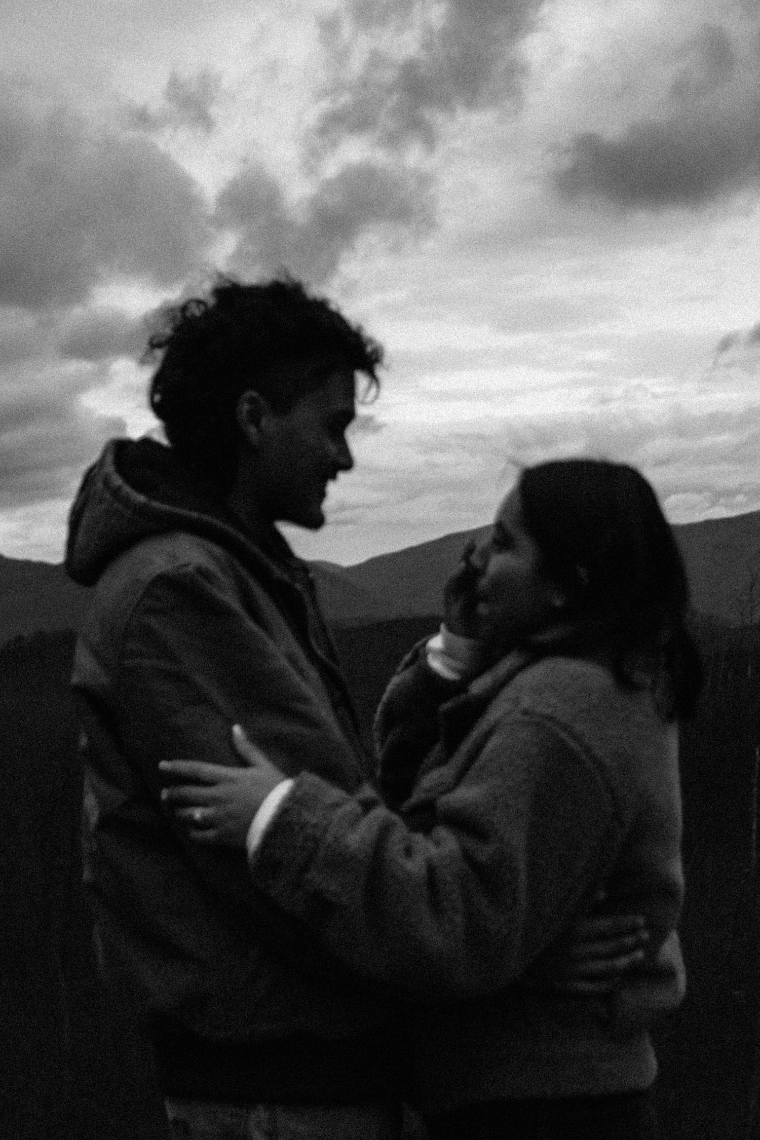 A romantic black and white moment shared between two people embracing against a cloudy mountain backdrop.